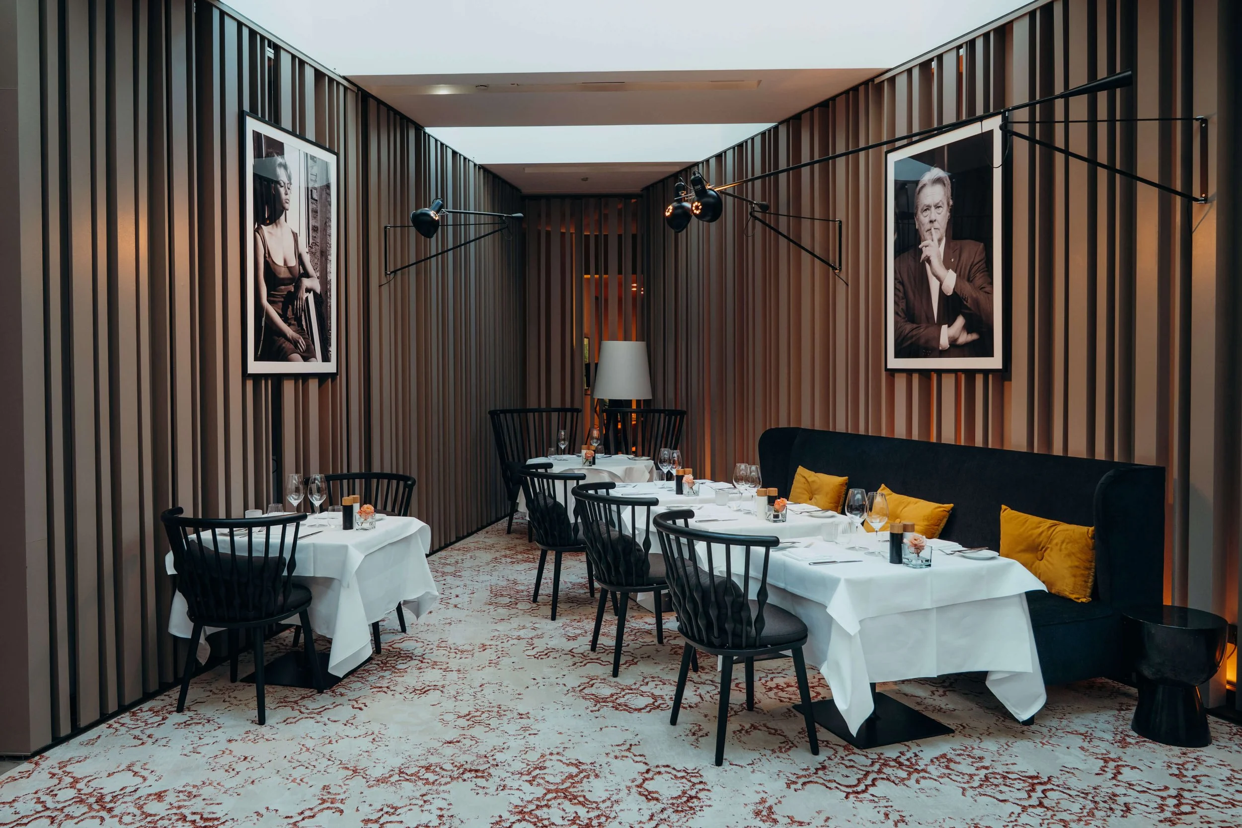 Elegant restaurant dining area with white tablecloths, black chairs, and framed black-and-white portraits on wooden paneled walls.