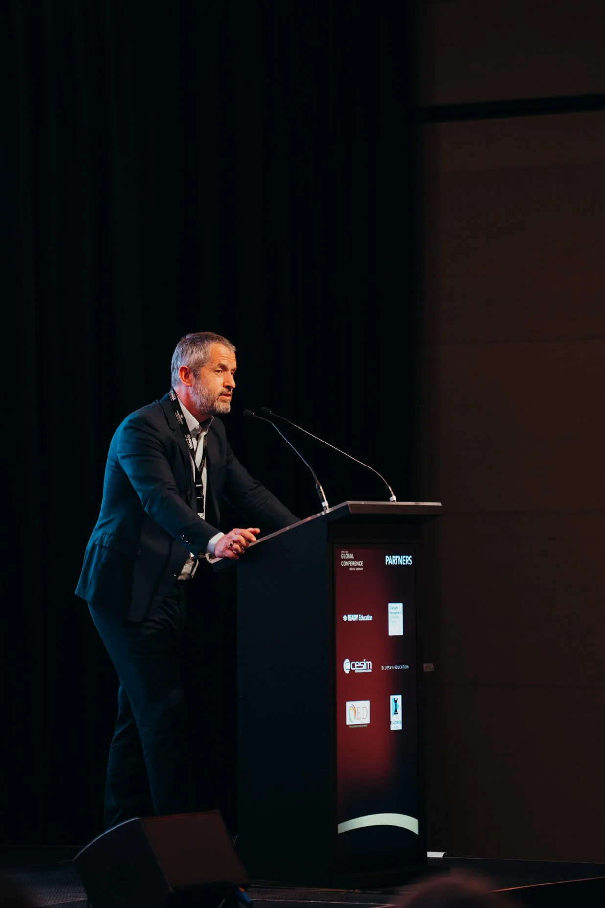 A man with gray hair and beard wearing a dark suit standing at a presentation podium with a microphone, speaking at a conference in a darkened room.