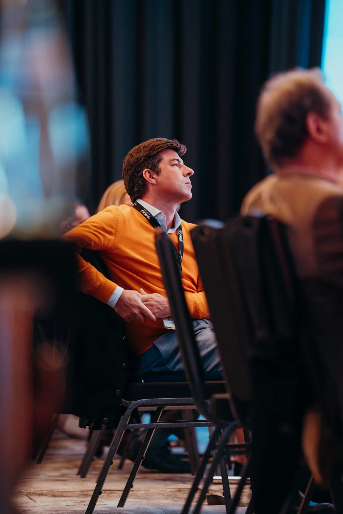 A man wearing an orange sweater and gray pants is sitting in a conference room, attentively listening, with a black lanyard around his neck.