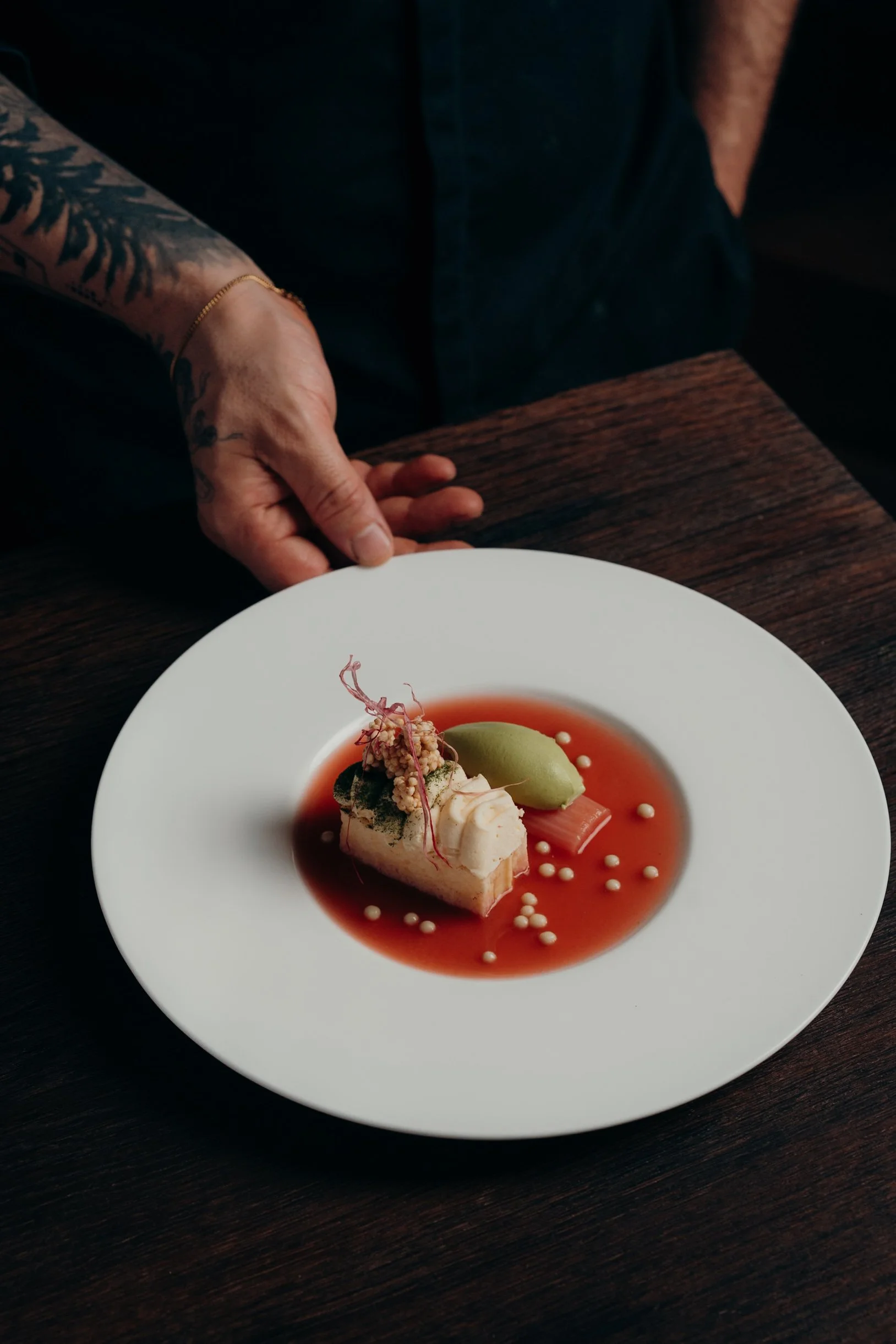 Ein Teller mit elegante Nachspeise, bestehend aus einer cremigen Komponente, einer grünen Kugel, roten Gelee und dezenten weißen Perlen, auf einem dunklen Holztablett.