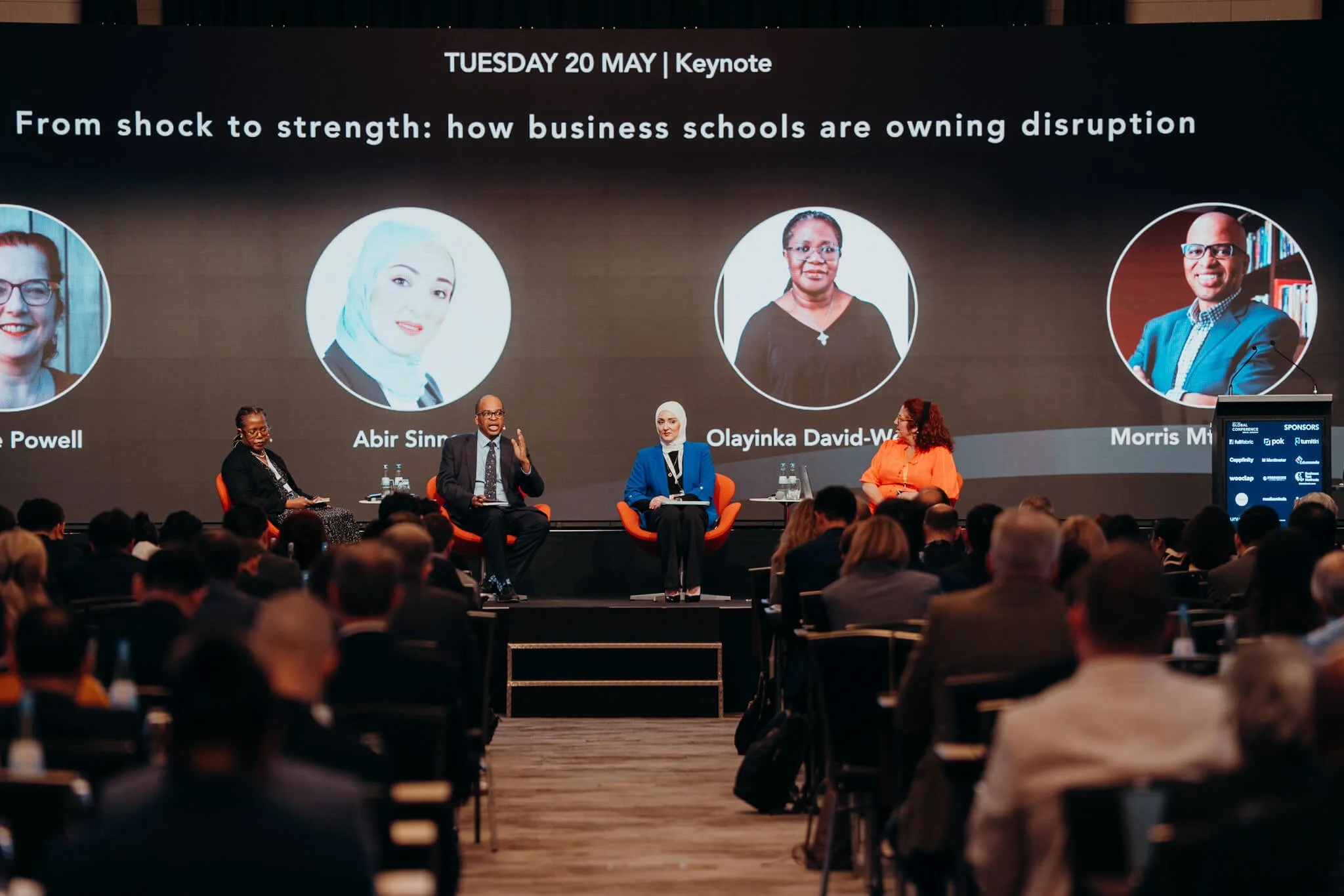 Panel discussion at a conference with four speakers seated on stage in front of a large screen displaying panelists' photos and names, audience listening.