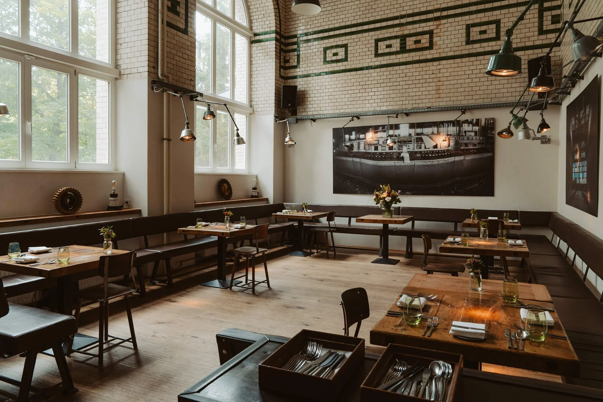 Empty restaurant with wooden tables and chairs, large windows, modern hanging lights, framed artwork on brick walls, and small centerpieces on tables.