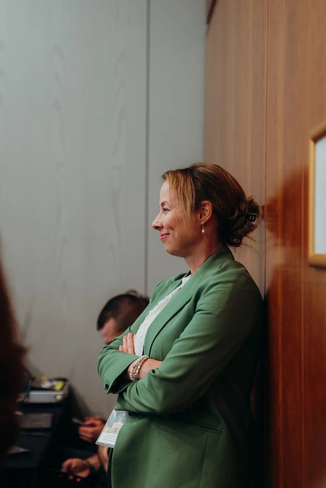 A woman in a green blazer with arms crossed, leaning against a wooden wall, smiling, in an indoor setting.