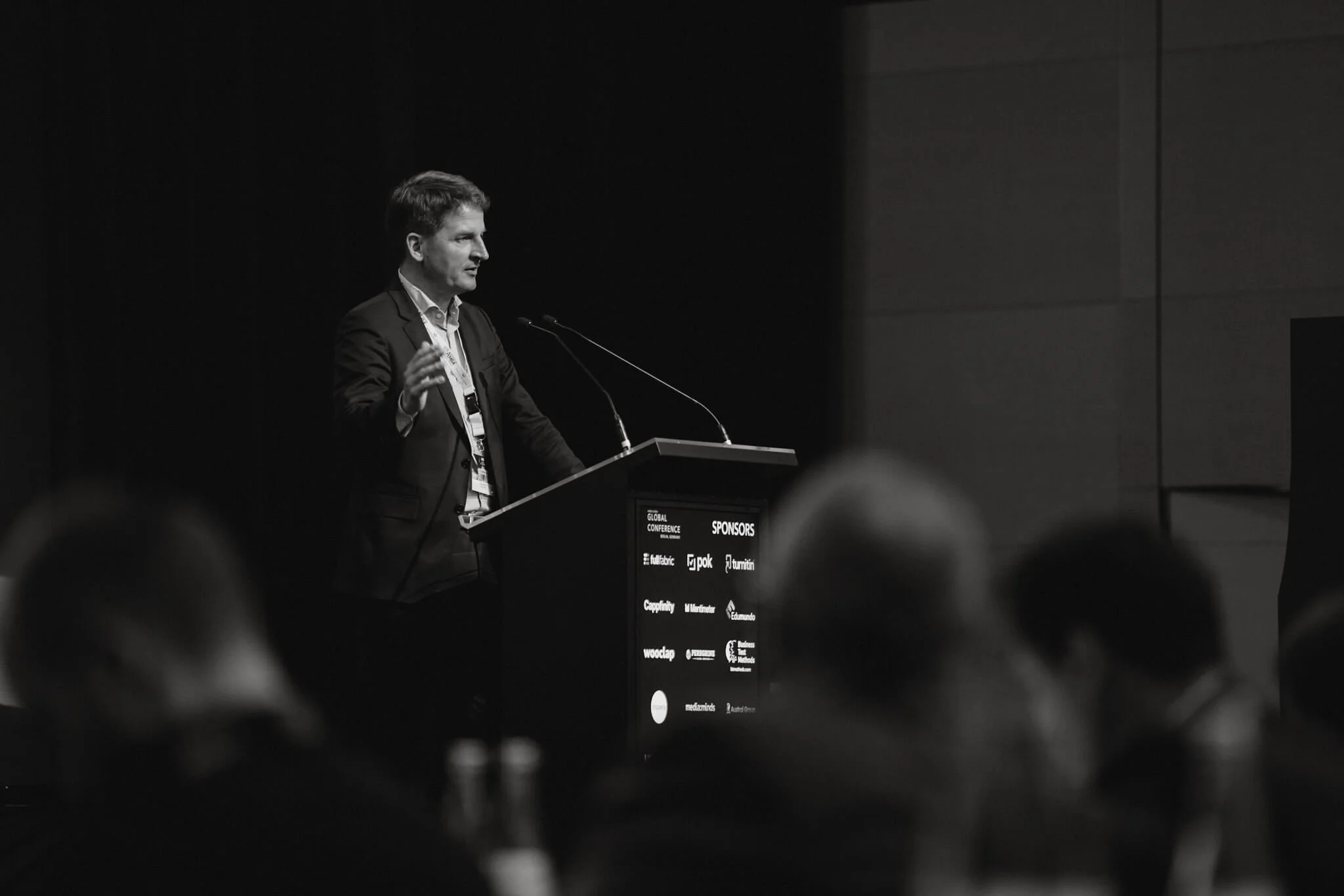 A man giving a presentation at a conference, standing at a podium with a microphone, in a dark room with an audience.