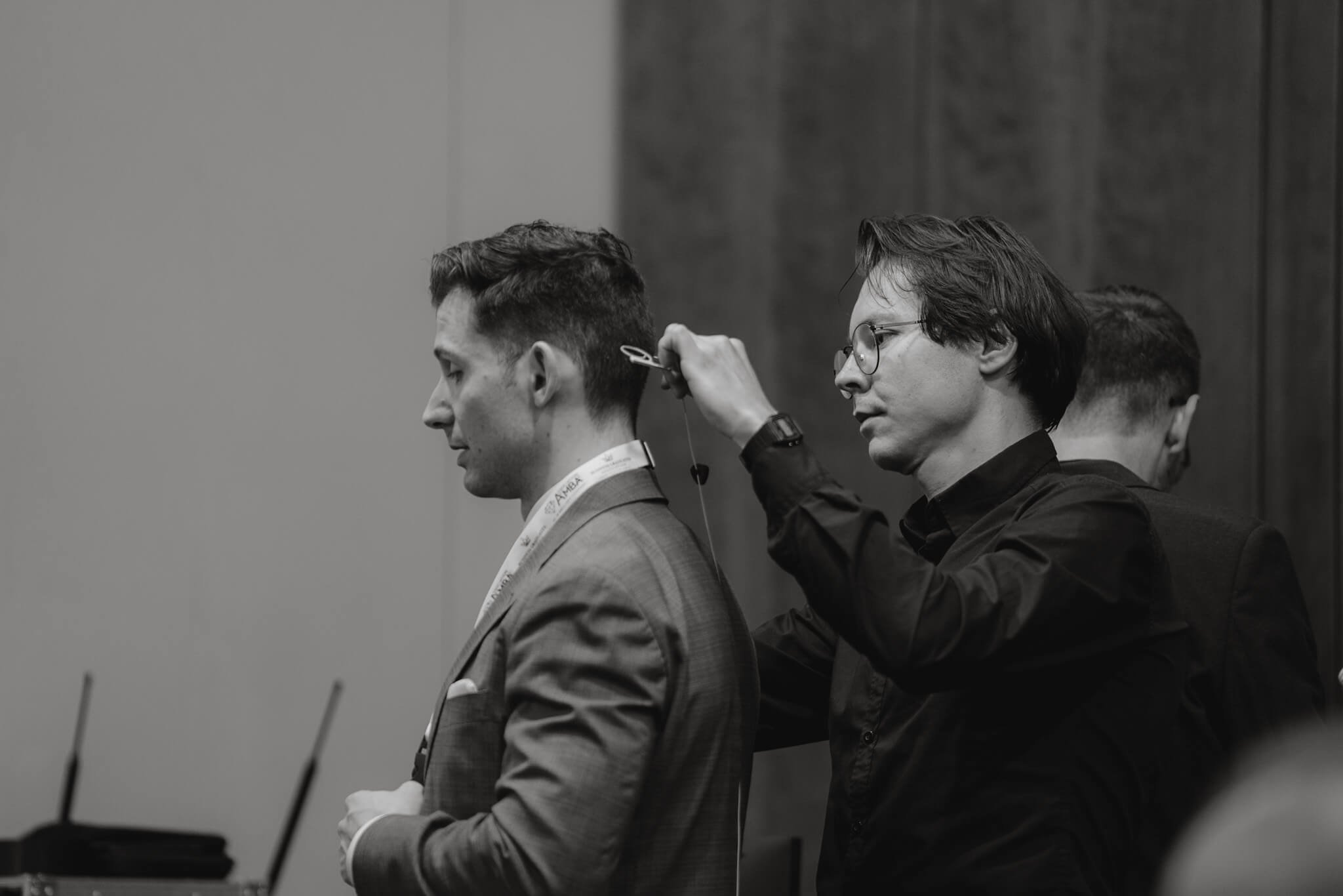 A man with glasses and long hair is adjusting a microphone on another man who is smiling and wearing a suit at a conference or event.