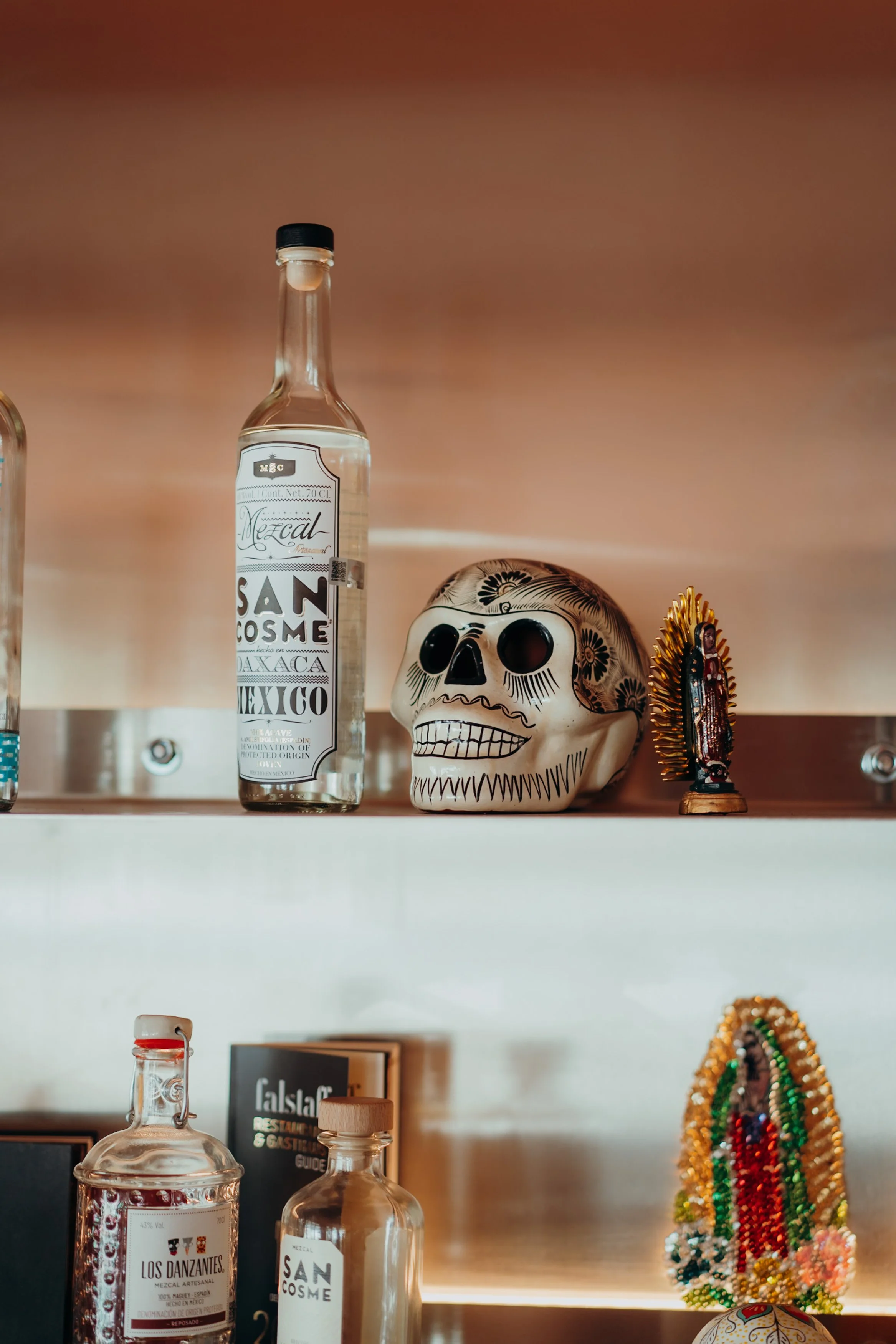Dekorative Gegenstände auf einer gleichfarbigen Wand. Darunter befinden sich einige Schnapsflaschen, eine weiße Totenkopf-Deko mit schwarzen Verzierungen, ein religiöser Schmuck und eine farbenfrohe Beads-Statue.