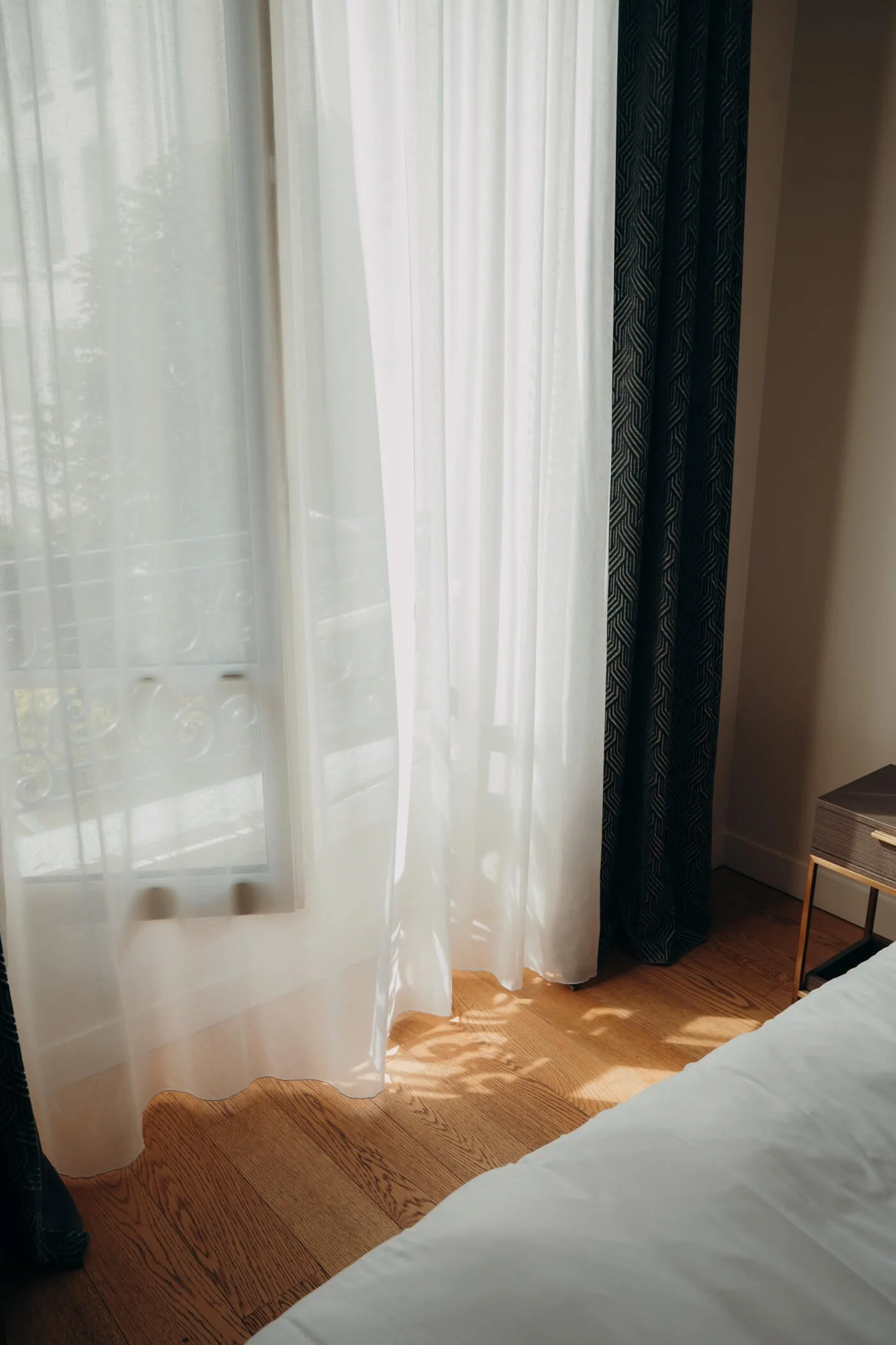 Blick auf einen Vorhang im Zimmer mit Holzfußboden, teilweise sichtbares Bett und Nachtisch. Außen ist ein Balkon mit Balkonreling sichtbar.