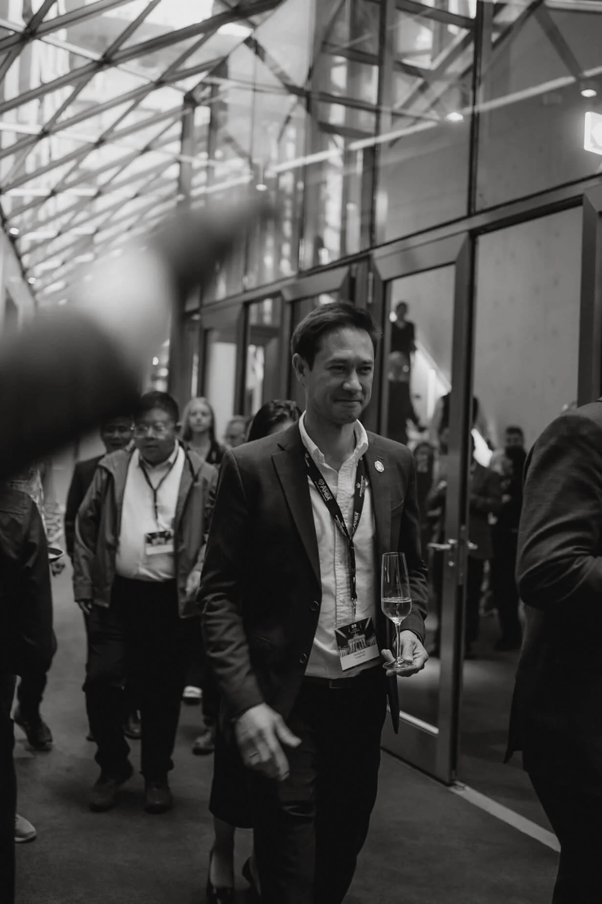 A man in a suit walking through a crowded indoor event, holding a glass of champagne, in a modern venue with glass walls and a high ceiling.