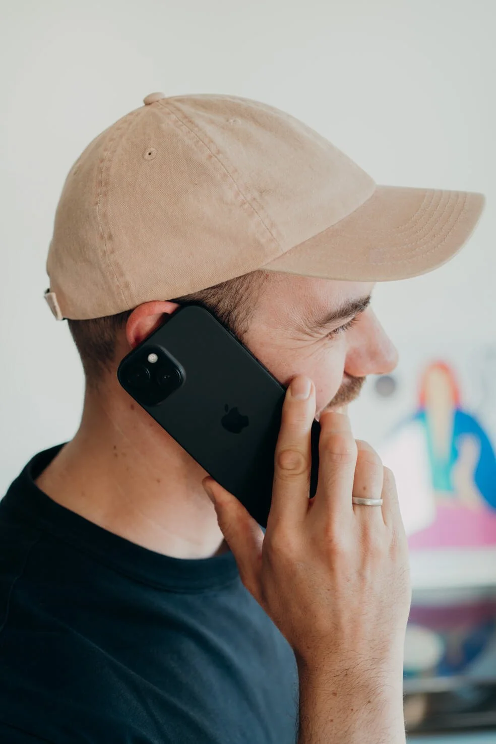 Ein Mann trägt eine beige Baseballkappe und spricht an einem schwarzen iPhone. Im Hintergrund ist eine bunte Illustration sichtbar.