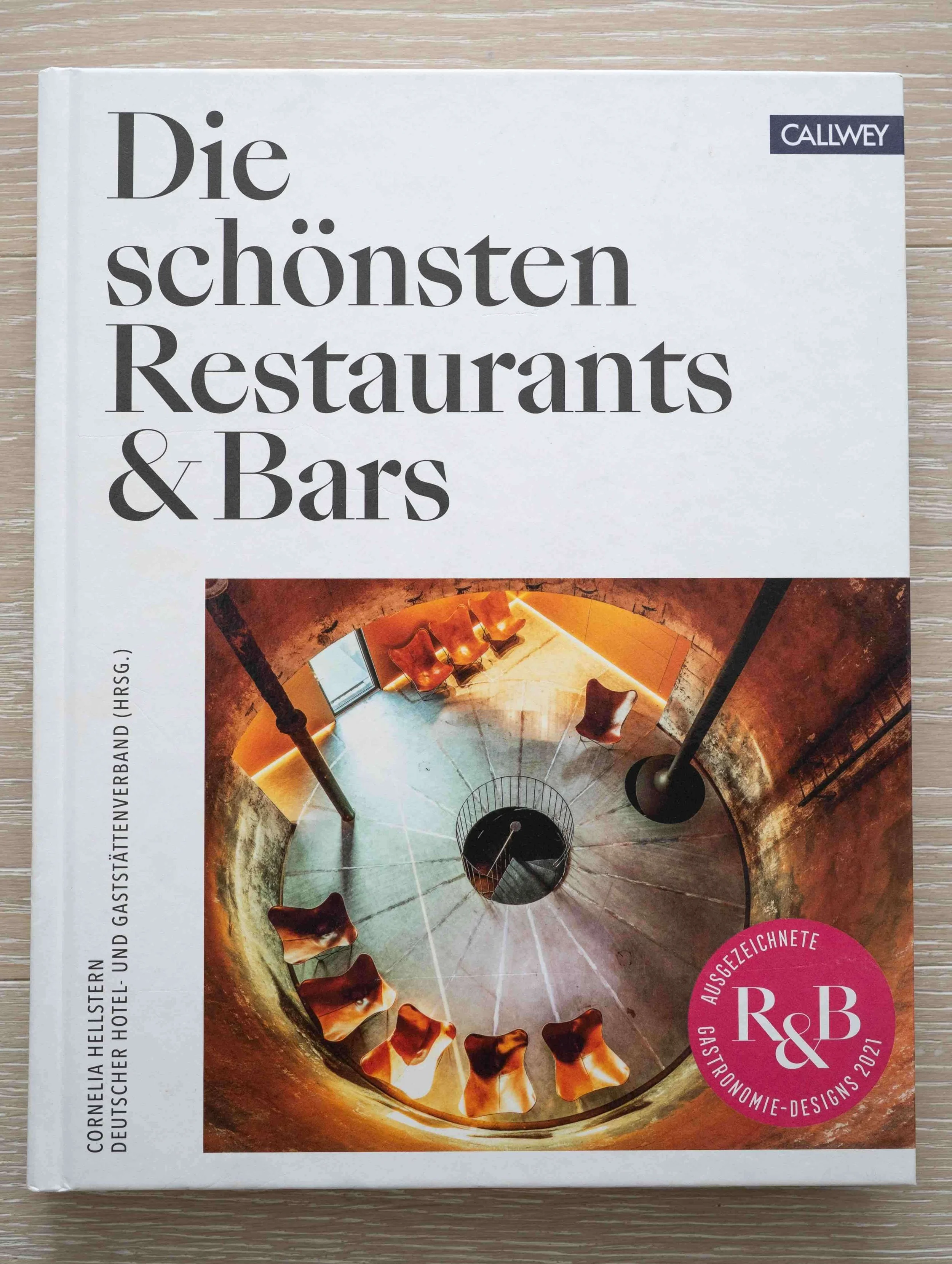 Buchcover mit Blick in einen kreisförmigen Schnitt durch einen Behälter mit Goldfischen, umgeben von roten und braunen Steinen, auf einem Holztisch.