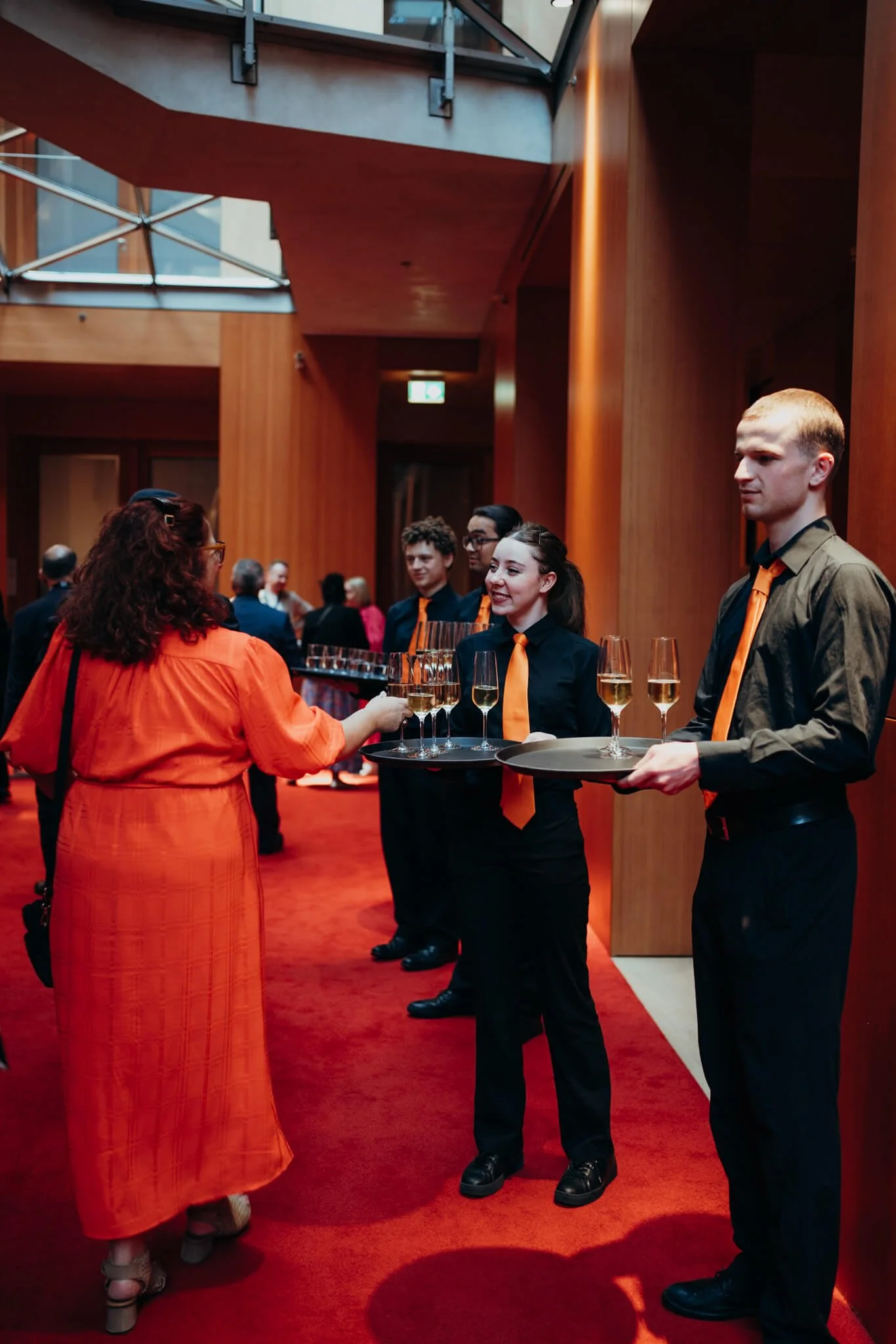 A woman in an orange dress is being served drinks by three waitstaff dressed in black shirts with orange ties at an indoor event with wooden walls and red carpet.
