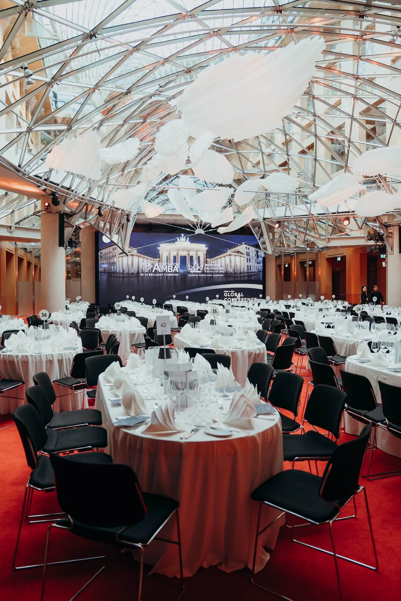 A conference hall decorated for a formal event with round tables covered with white tablecloths, neatly folded napkins, glassware, and silverware. The hall has a high, glass-domed ceiling with white cloud-like decorations hanging from it. The room fe