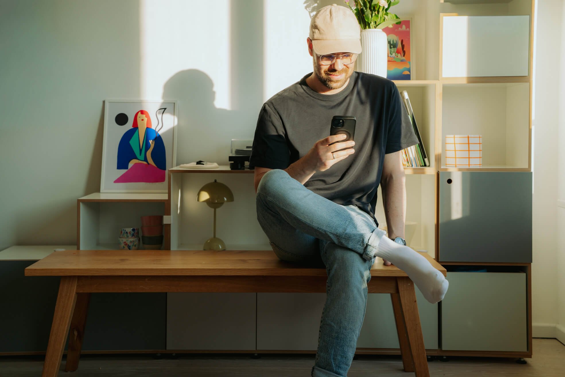 Ein Mann sitzt auf einem Tisch in einem modernen Wohnzimmer, trägt eine Baseballkappe, Brille und bequeme Kleidung, hält ein Smartphone in der Hand und lächelt. Im Hintergrund sind Bücherregale, eine Vase mit Blumen, ein Kunstbild und eine Lampe sichtbar.