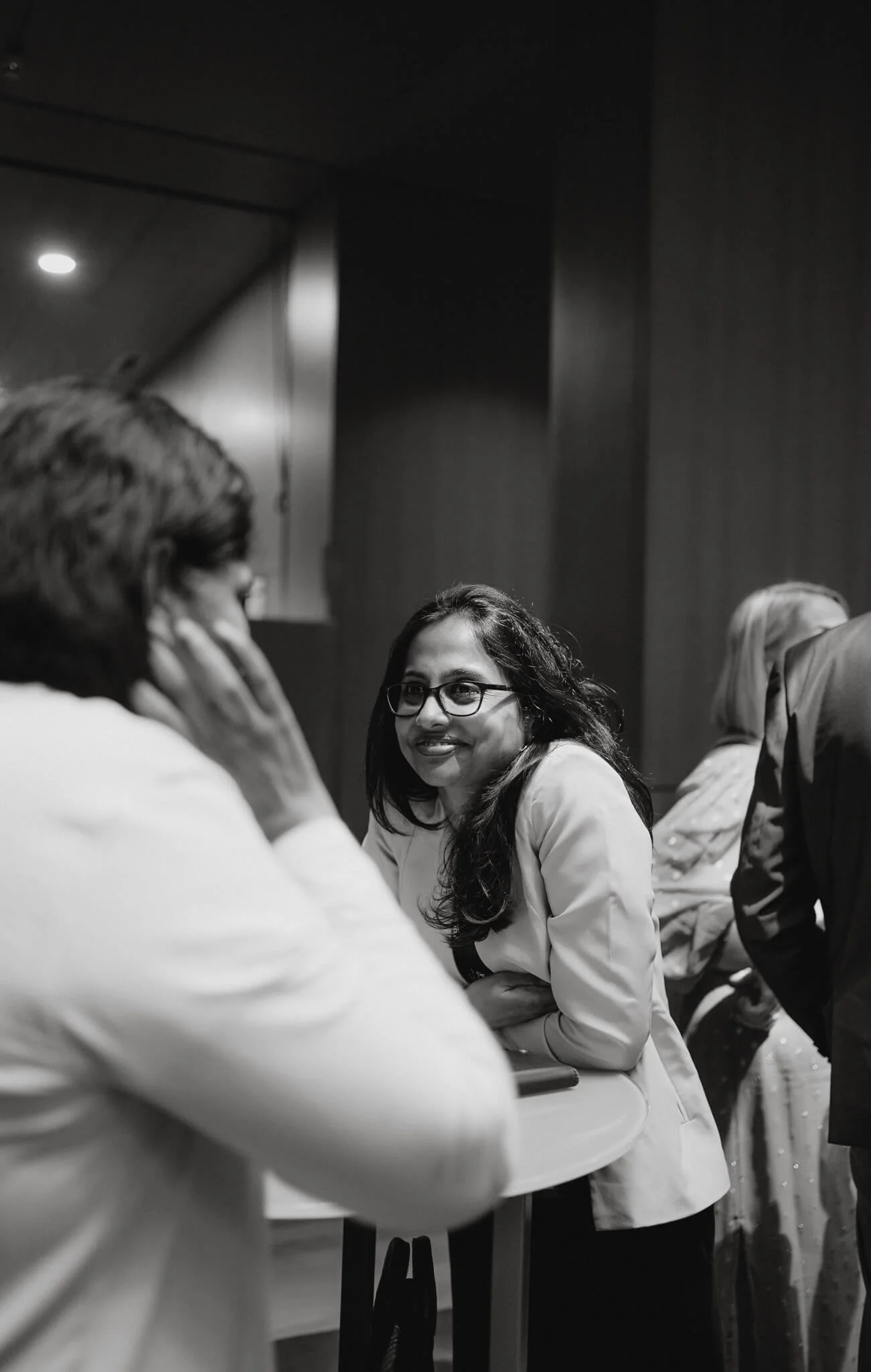 Black and white photo of a woman with glasses talking to two people at a table in a dimly lit room.
