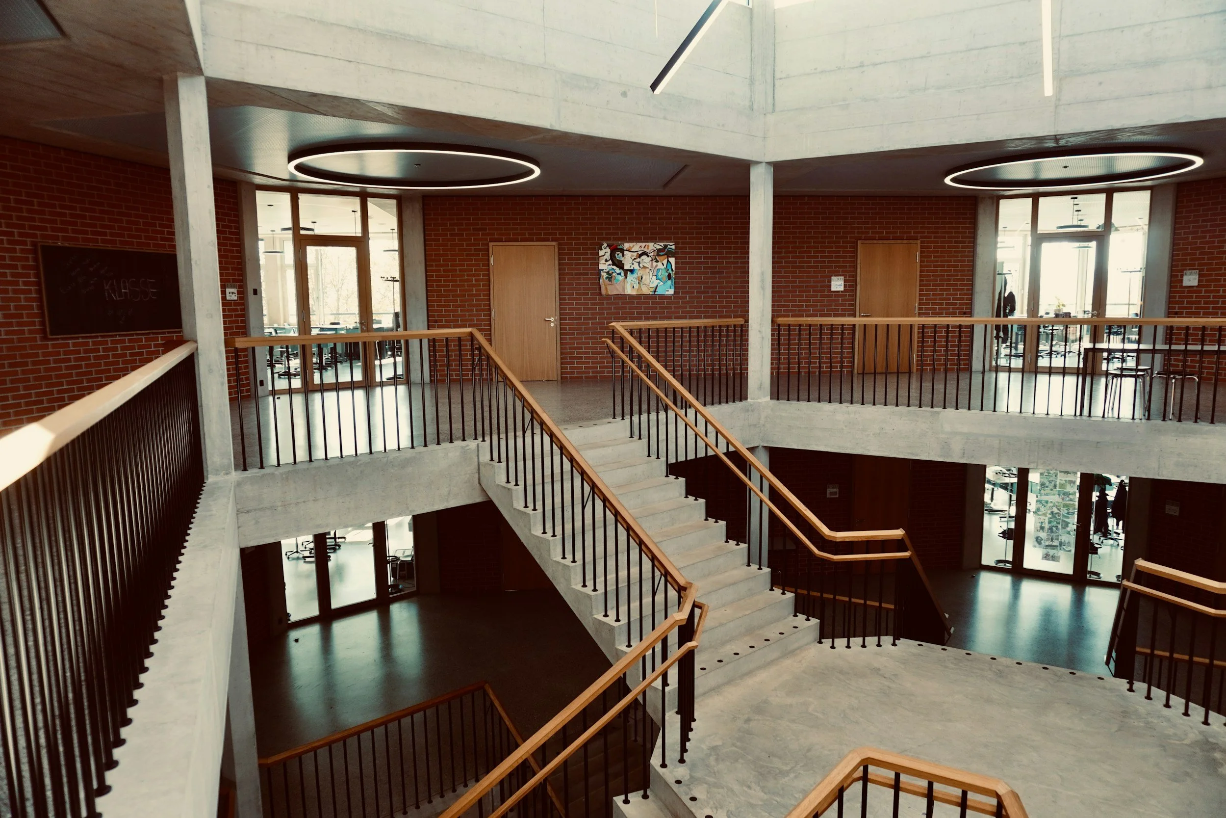 Interior of a modern multi-story building with concrete stairs, wooden handrails, and brick walls, illuminated by circular ceiling lights.