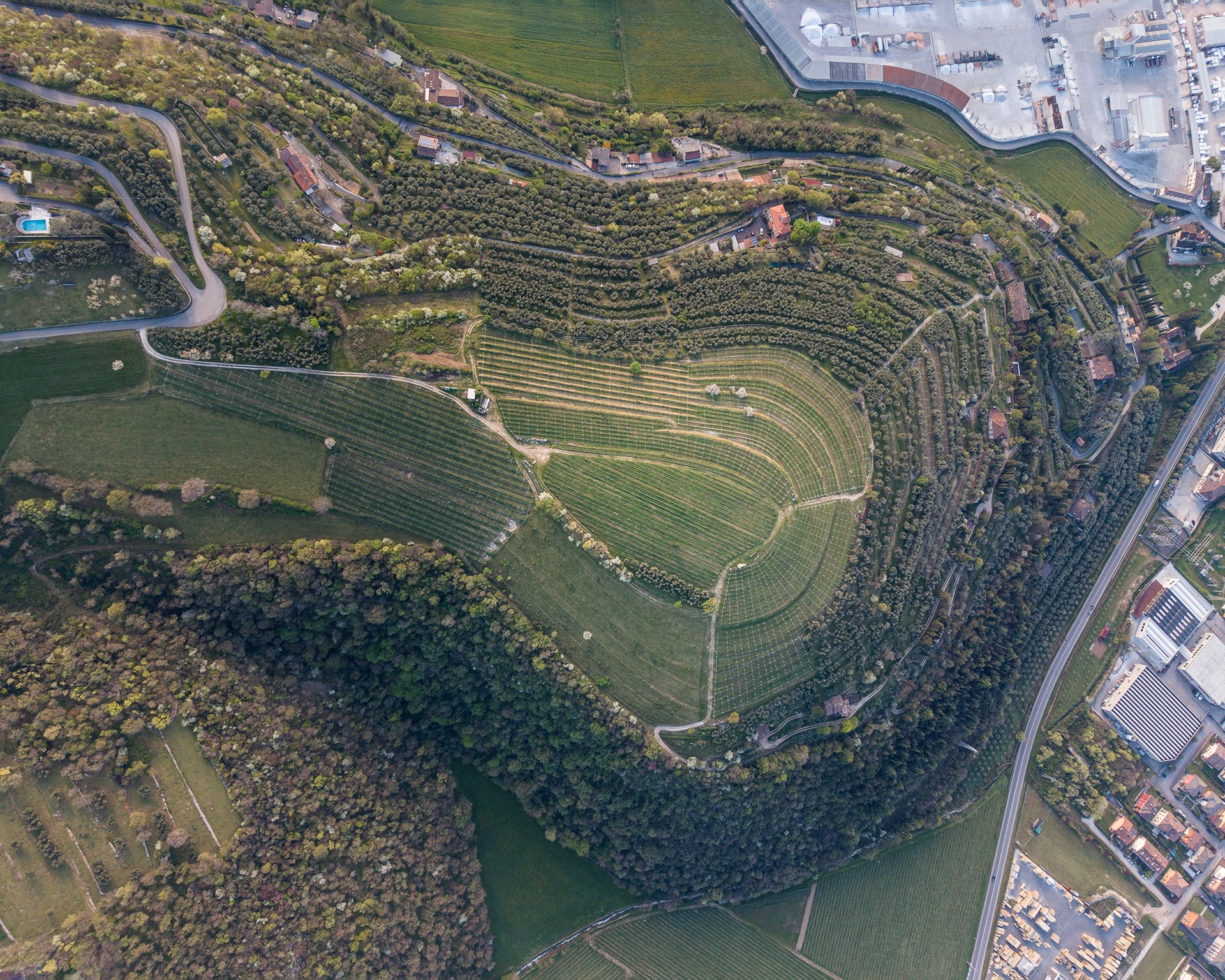 Immagine aerea di un paesaggio collinare con terreni agricoli, campi coltivati, alberi, alcune case e strade che attraversano il paesaggio.