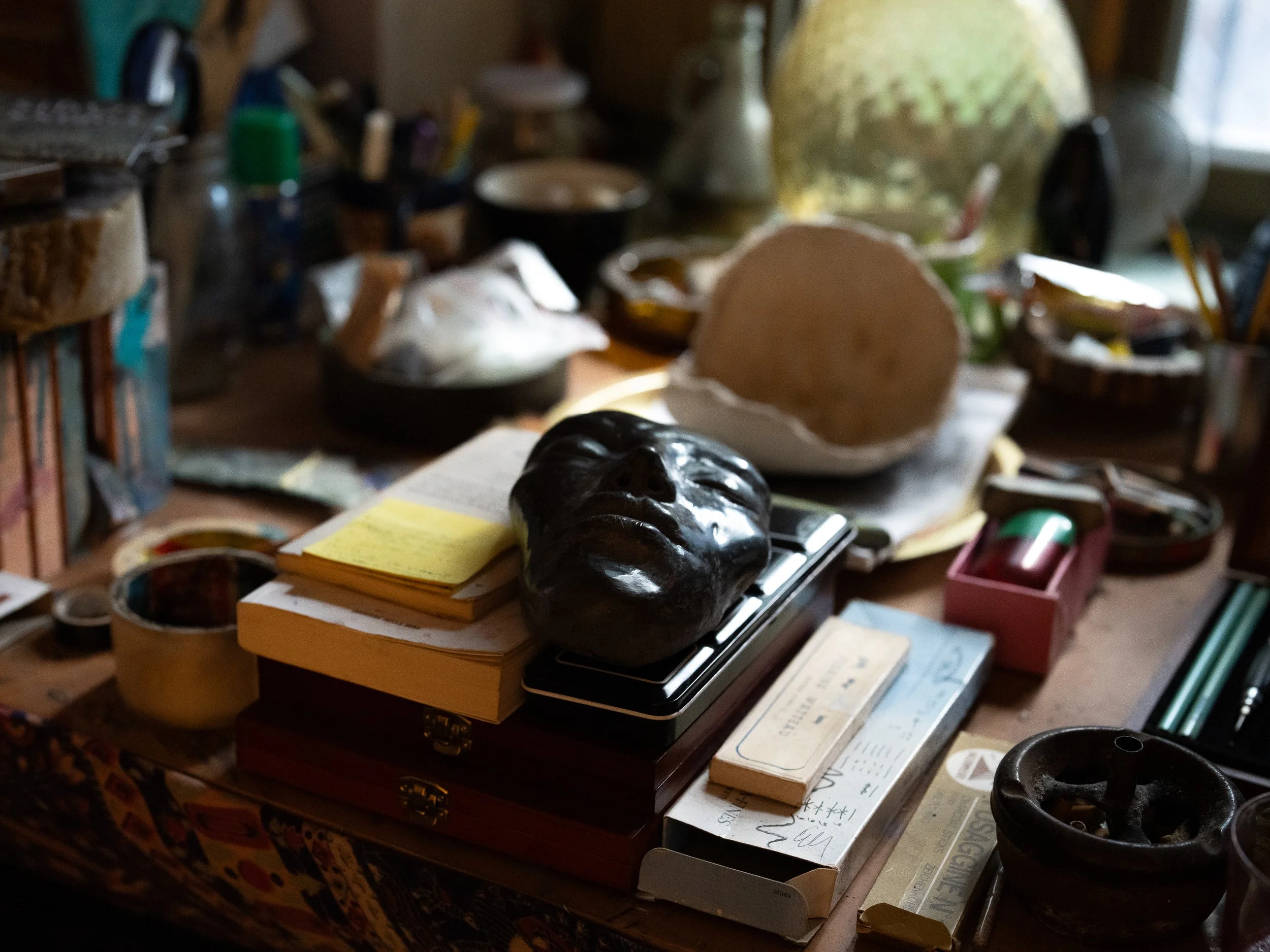 Una scrivania disordinata con vari oggetti tra cui libri, scatole, una maschera nera e altri oggetti vari. L'ambiente è scarsamente illuminato e sembra essere un'area di lavoro o di studio casuale.
