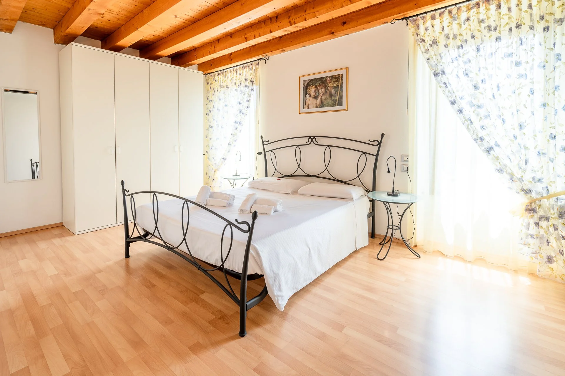 Camera da letto con letto matrimoniale, finestra con tende floreali, quadro sulla parete, candelabri, armadio bianco e pavimento in legno.