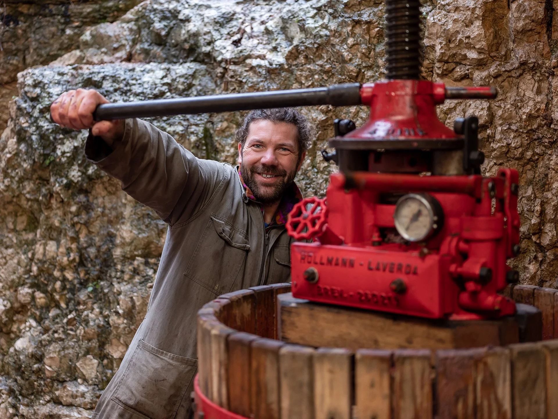 Uomo con barba e capelli ricci, indossa una giacca marrone, mentre utilizza una pressa idraulica rossa in un'area naturale rocciosa.