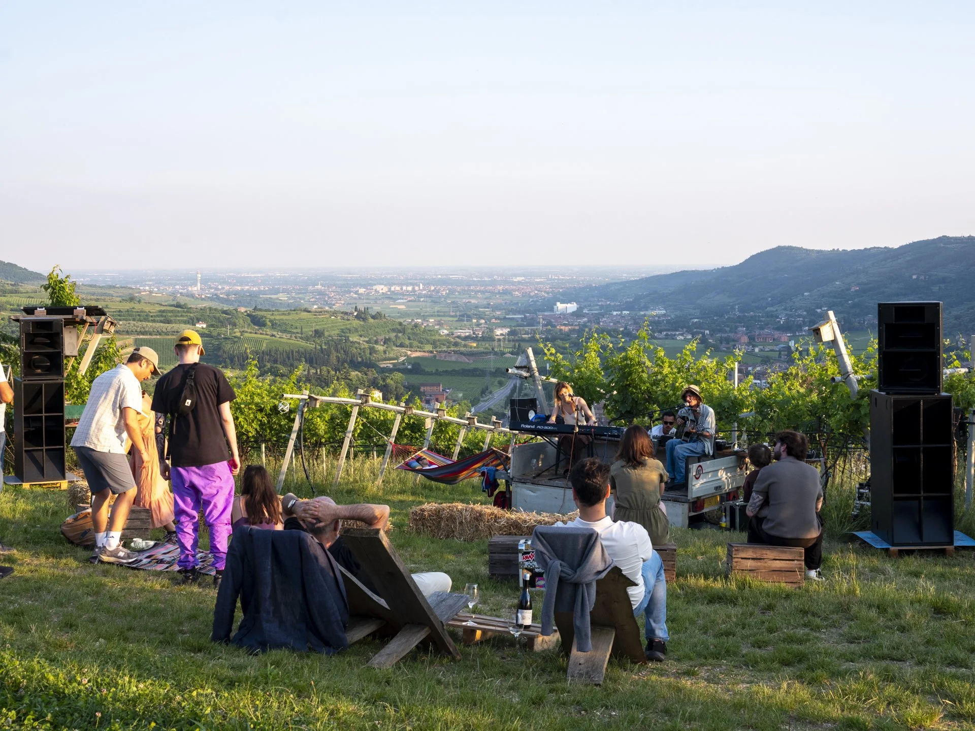 Gruppo di persone che si riuniscono in un vigneto con vista sulla campagna e le montagne, mentre ascoltano un concerto dal vivo con strumenti musicali, come tastiere e chitarre, e casse audio.
