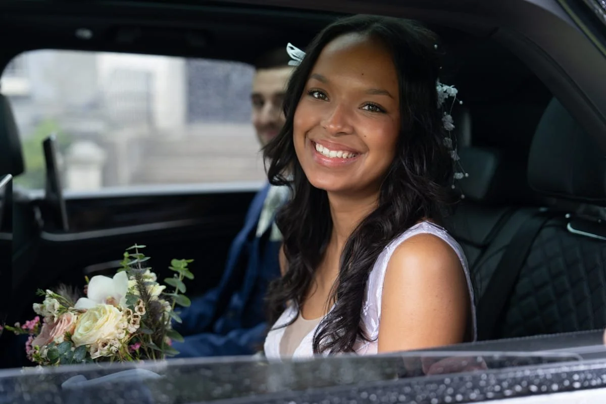Una donna con acconciatura decorativa con fiori, che sorride seduta in una macchina con un uomo sullo sfondo. La donna tiene un bouquet di fiori.