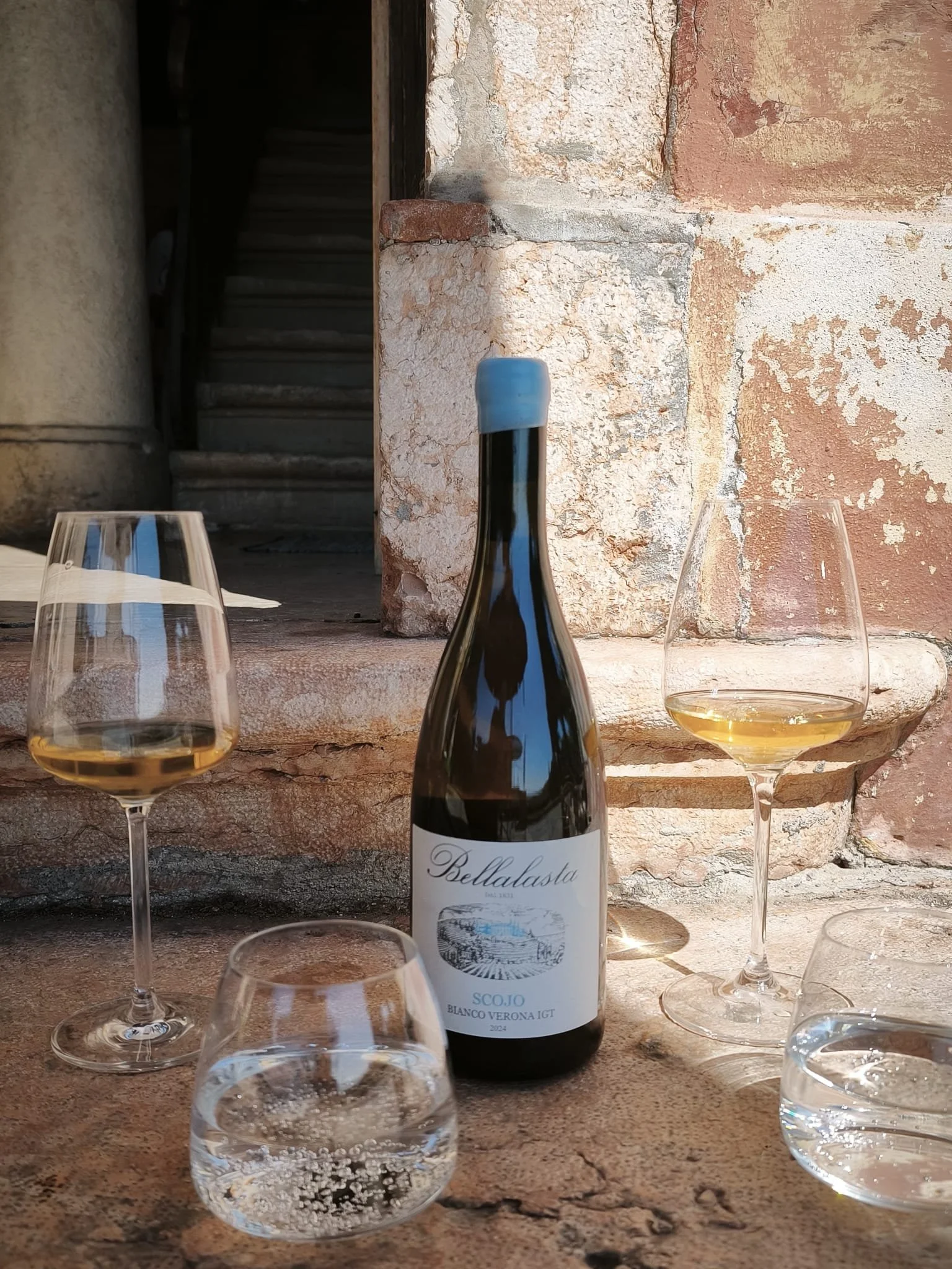 Una bottiglia di vino bianco con il nome 'Bellavista' e due bicchieri di vino bianco sono posti su un tavolo di pietra. Sullo sfondo, un muro di pietra e una scala che porta al piano superiore.