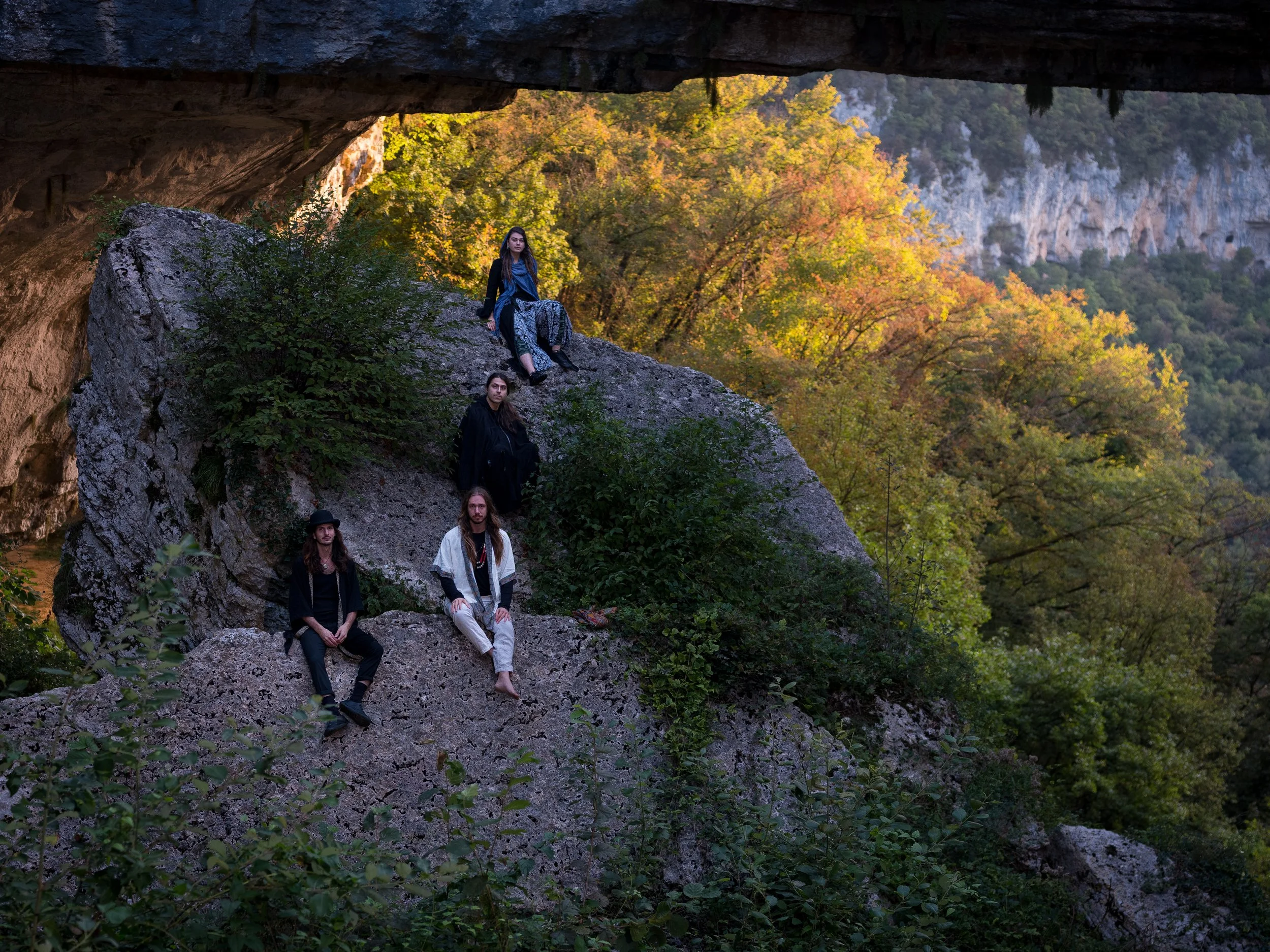 Cinque persone sedute su rocce sotto un ponte in una foresta con foglie autunnali, in un paesaggio naturale.