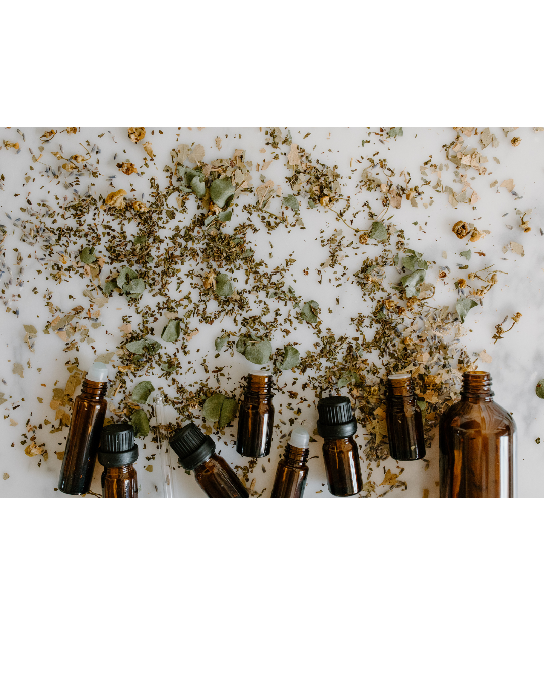 Multiple small amber glass bottles with black and white caps on a white surface surrounded by scattered dried herbs and leaves.