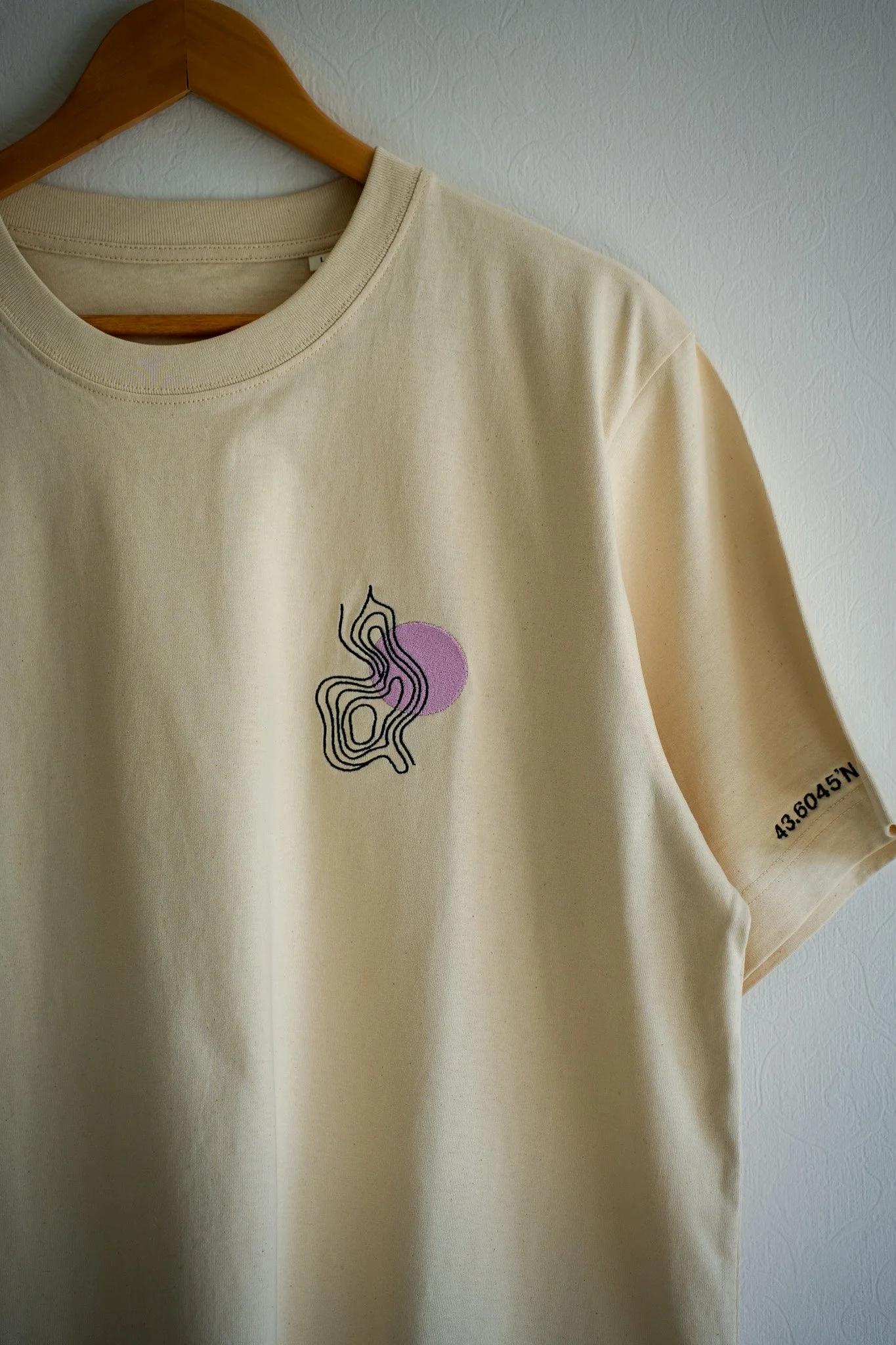 Cream-colored T-shirt hanging on a wooden hanger with a black abstract line drawing, a purple circle, and black text on the right sleeve showing coordinates.