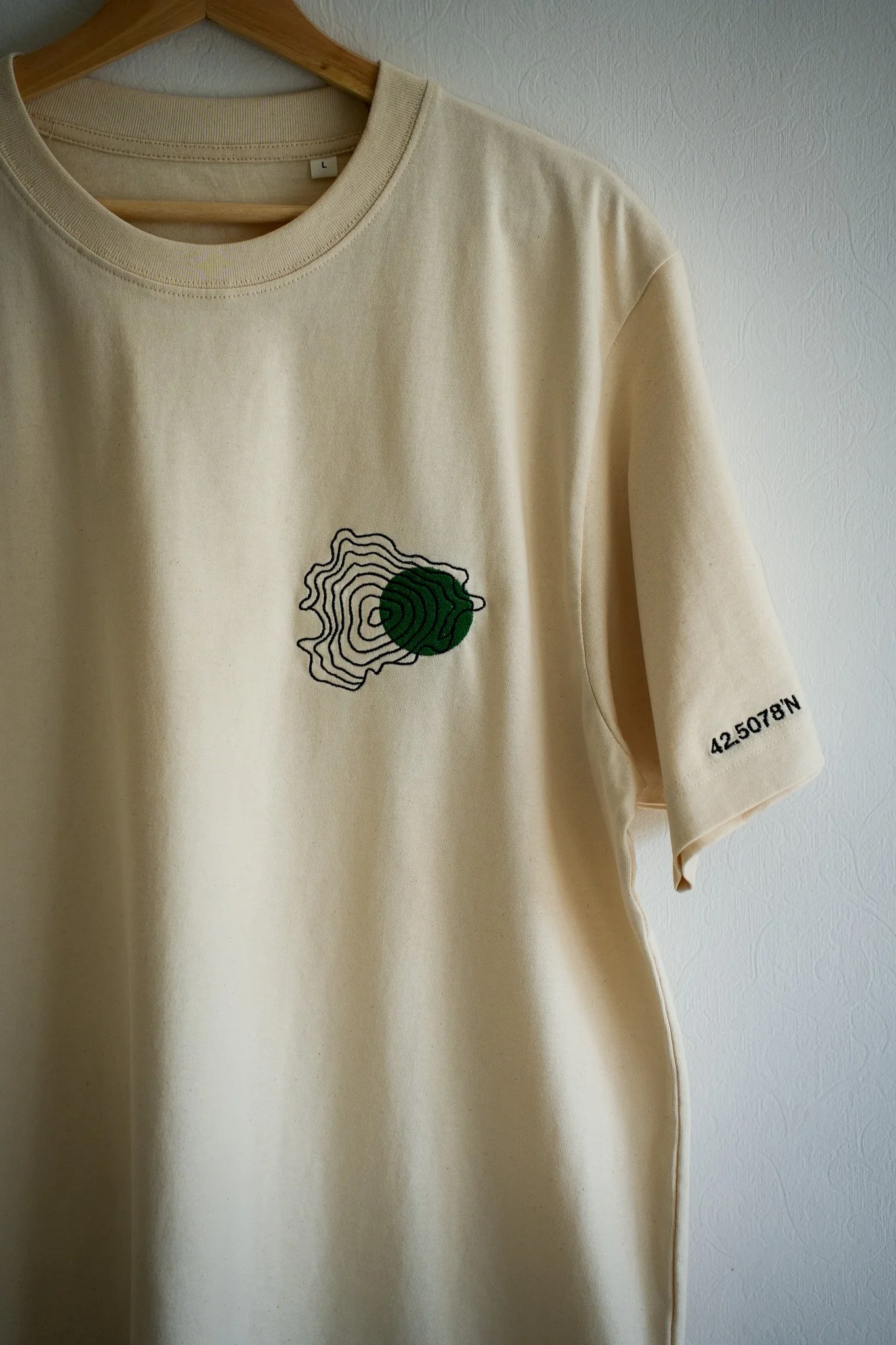Cream-colored t-shirt hanging on a wooden hanger against a white textured wall, featuring a black and green embroidered topographic map design on the front and a small black text '42 5078N' on the sleeve.