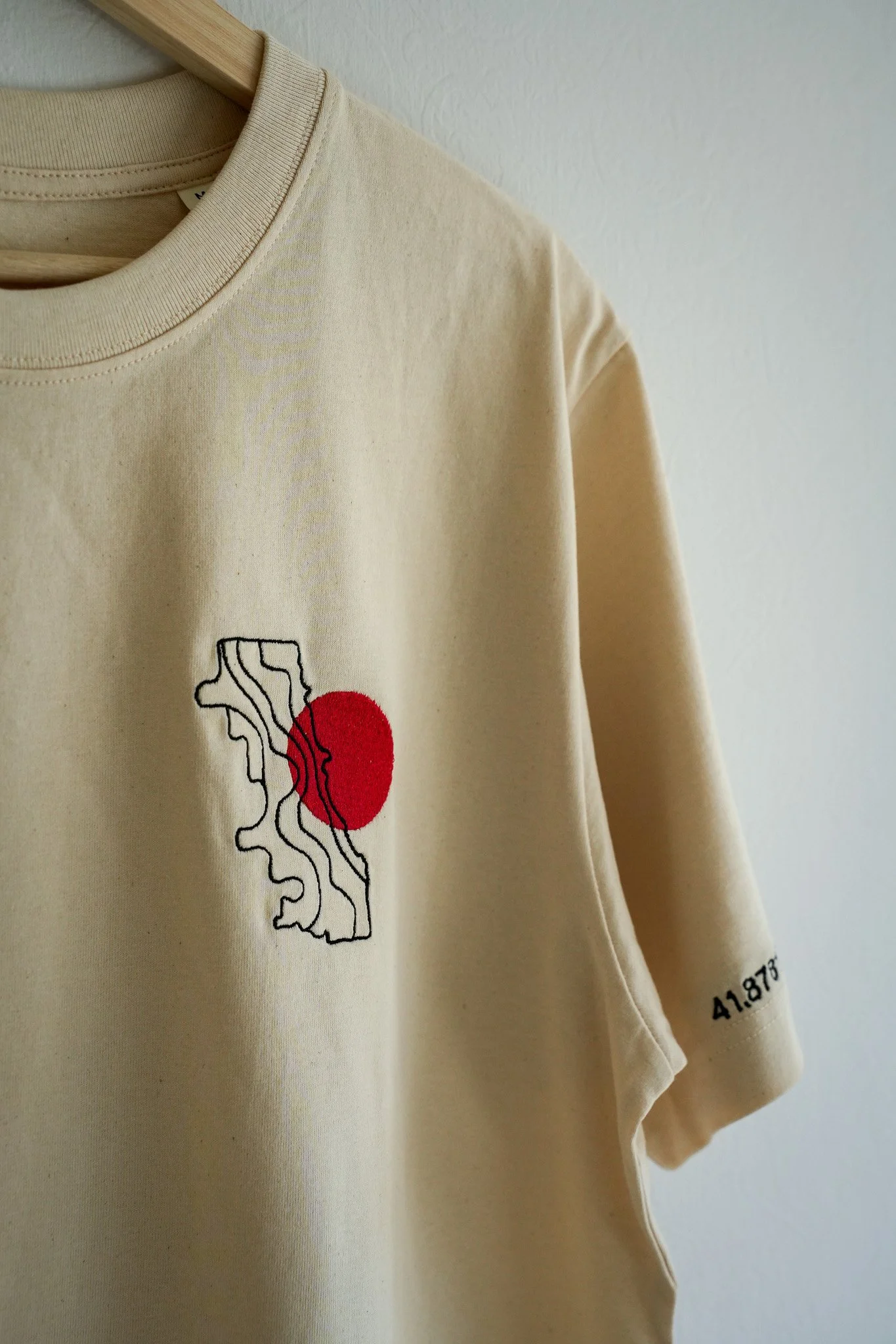 A beige T-shirt hanging on a wooden hanger, featuring a black line drawing of a map with a red circle in the center. The sleeve shows the coordinates 41.873.