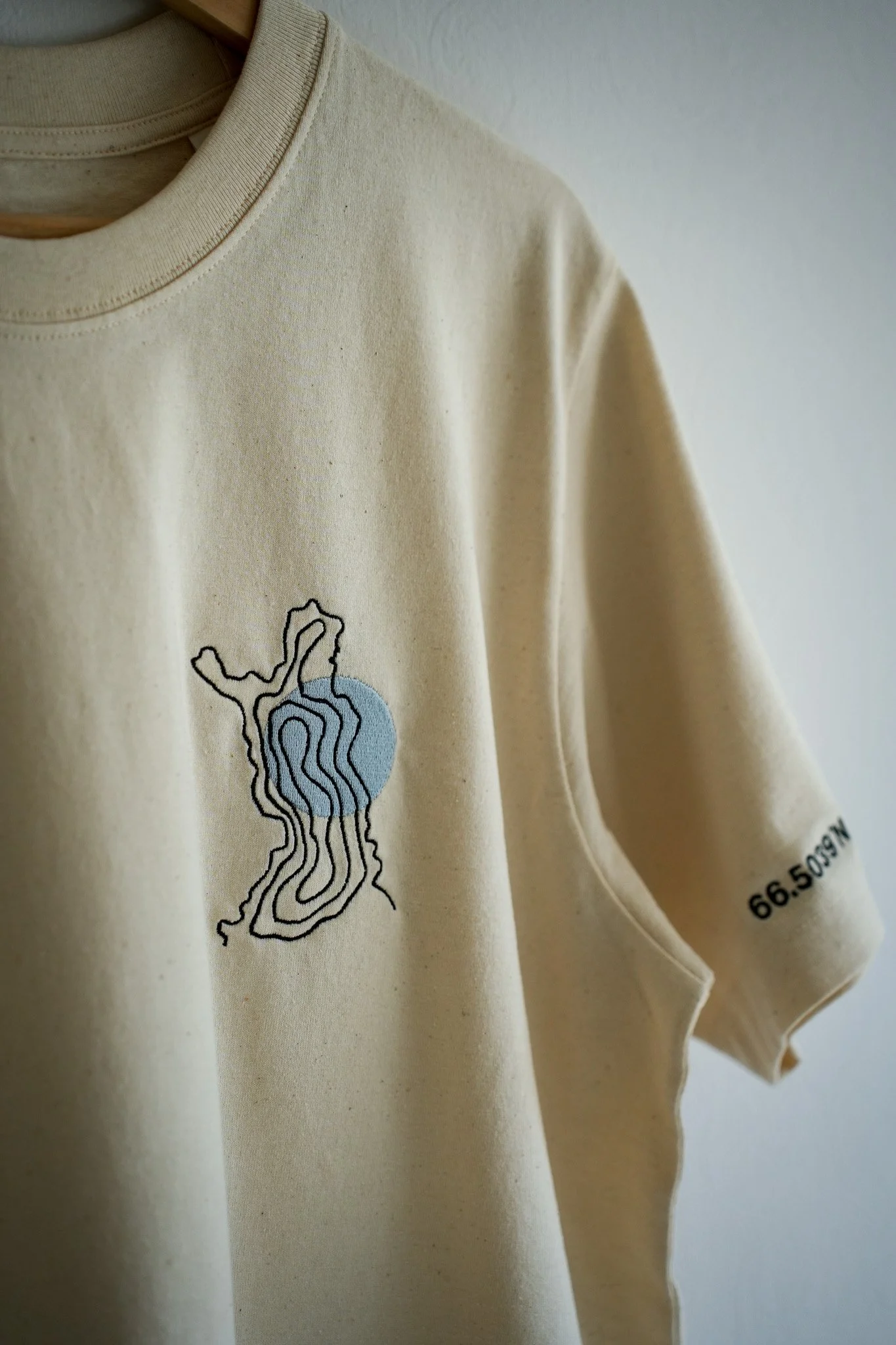 A beige T-shirt hanging on a wooden hanger, featuring a topographical map design with a light blue circle and black contour lines, and black text on the sleeve reading '66.509N'.