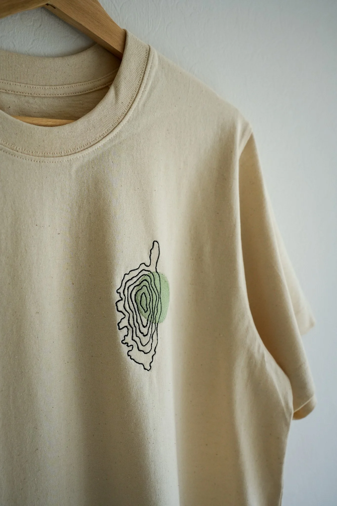 Beige T-shirt with black outline topographic map design and green highlight on front, hanging on a wooden hanger.