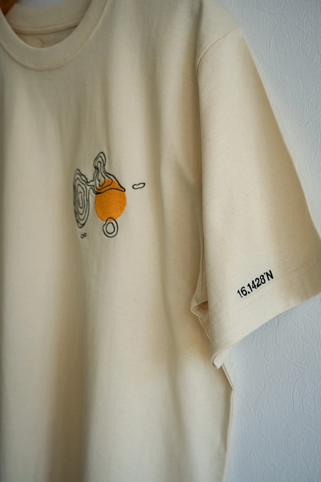 A beige T-shirt with a line drawing of a planet and orange circle on the front. The T-shirt has a black alphanumeric code 16I1428'N on the sleeve.