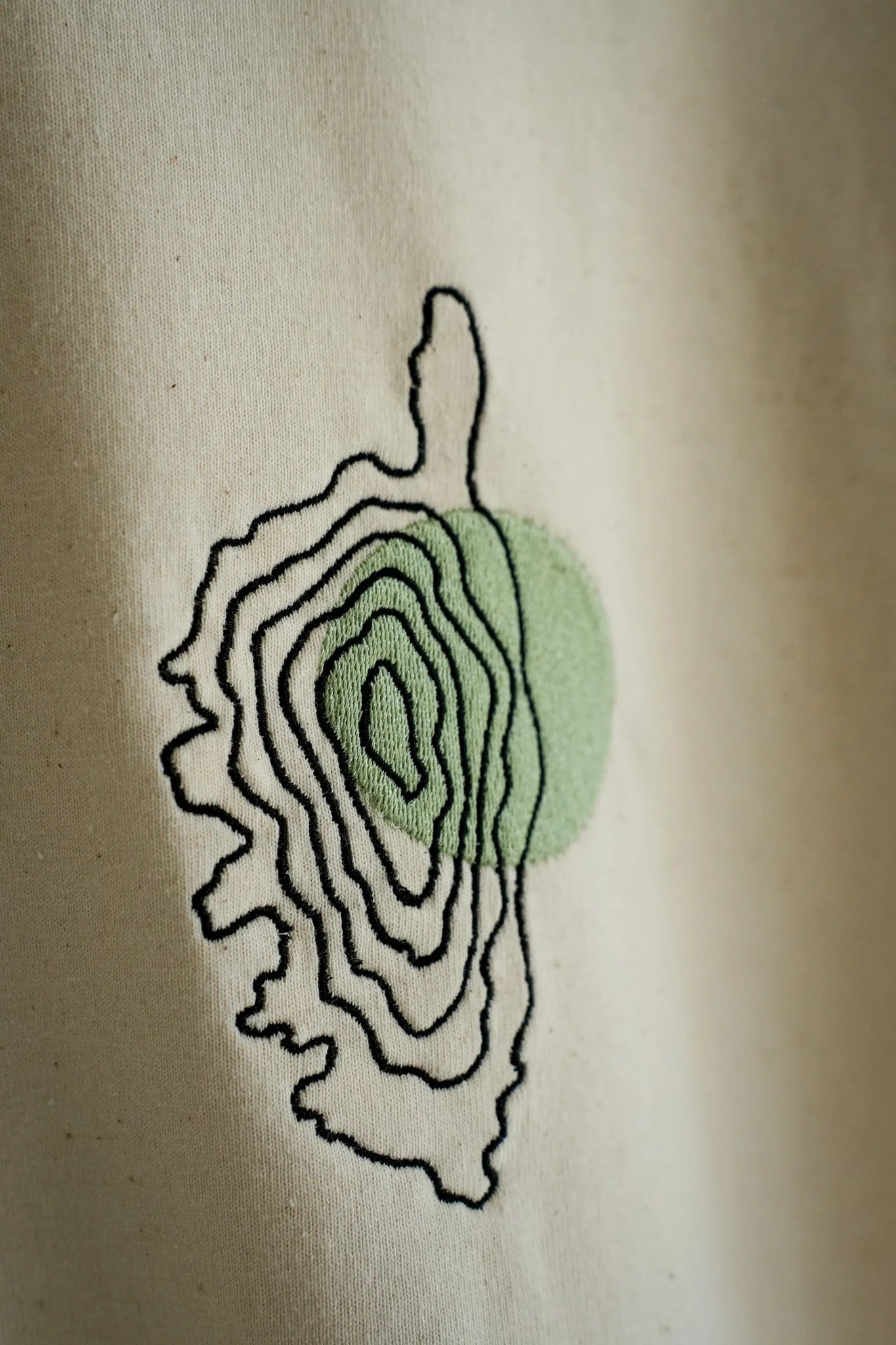 Close-up of embroidered design on fabric, featuring black contour lines and a green shape, resembling a topographical map.