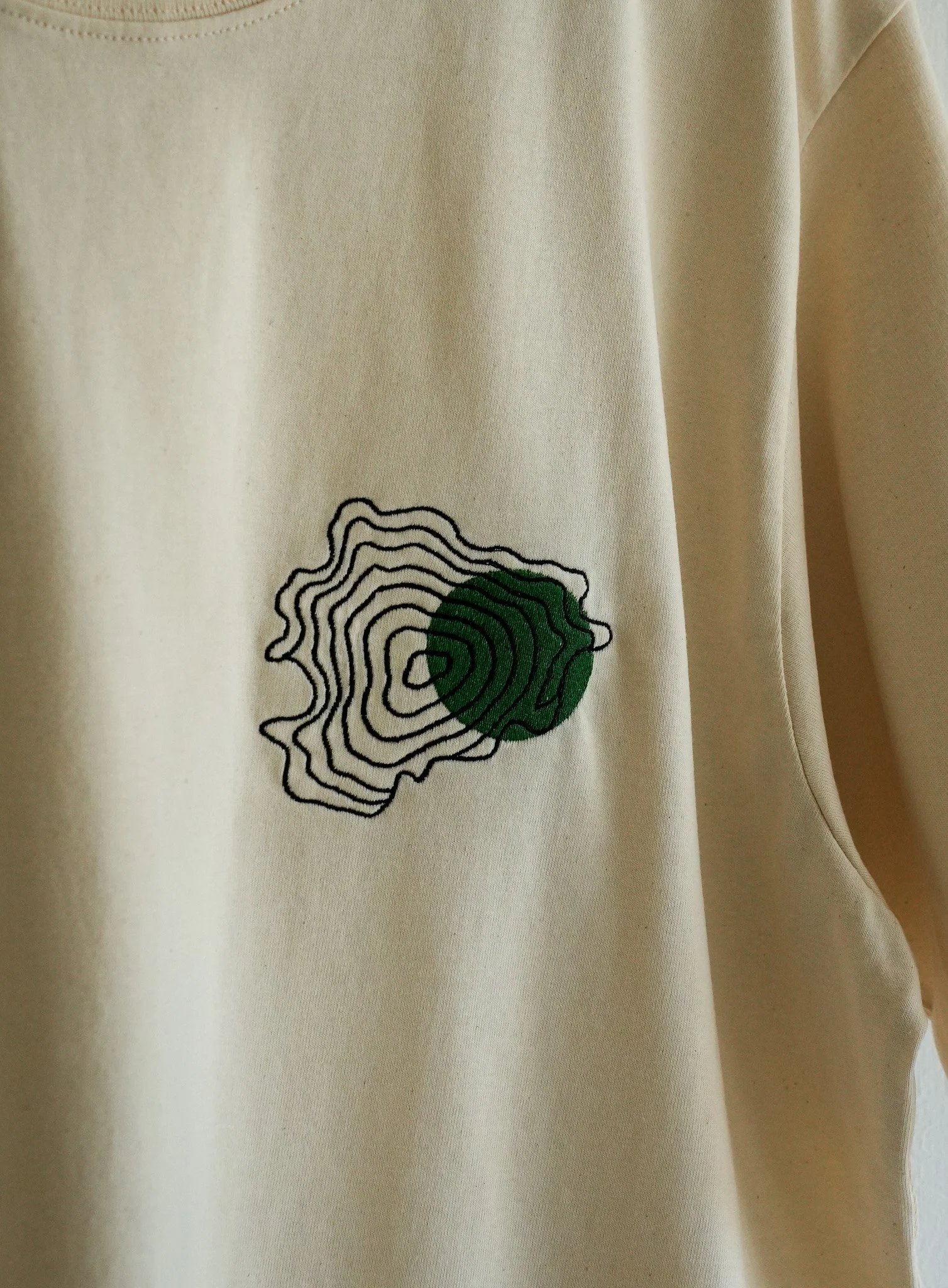 A beige shirt with a black and green embroidered design resembling a topographic map with a green shaded area.