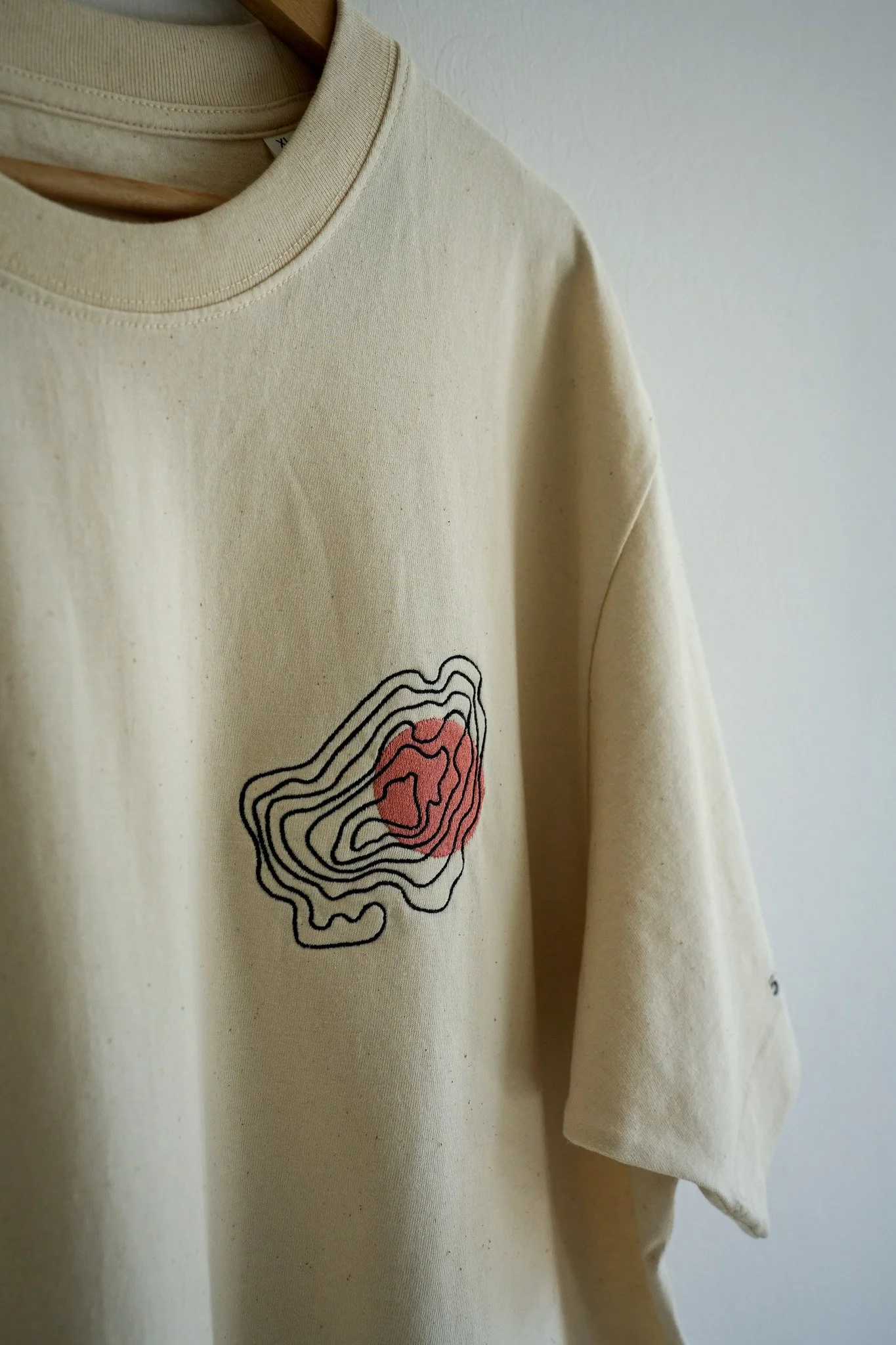Beige sweatshirt with a topographic map graphic featuring black contour lines and a red area, hanging against a white wall.