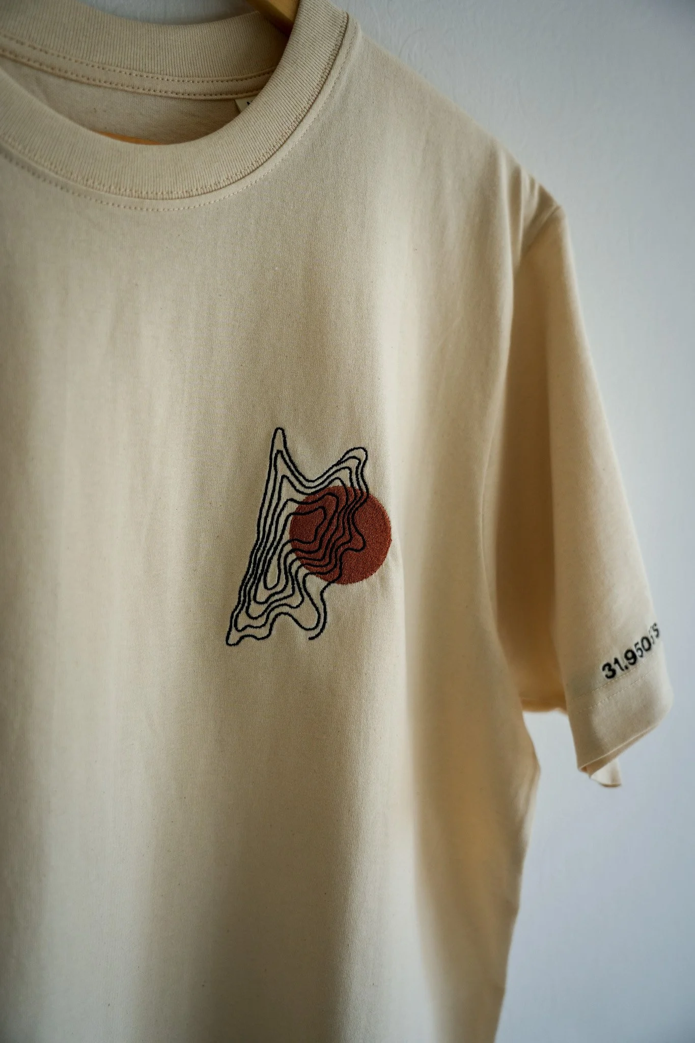 Cream-colored T-shirt with a black abstract line design and a red circle on the front, and small black text on the sleeve.