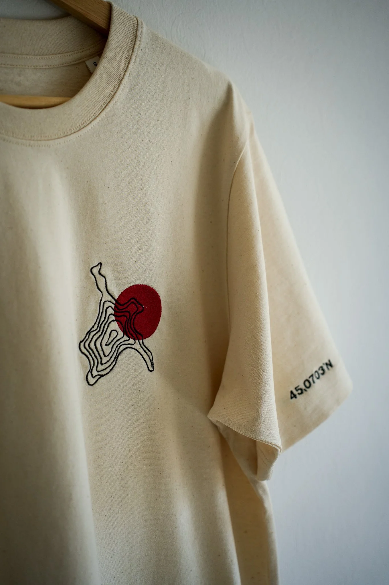 A beige T-shirt hanging on a wooden hanger. The T-shirt has a black line drawing of a topographic map with a red circle behind it near the chest area, and the coordinates 45.0703 N printed on the sleeve.