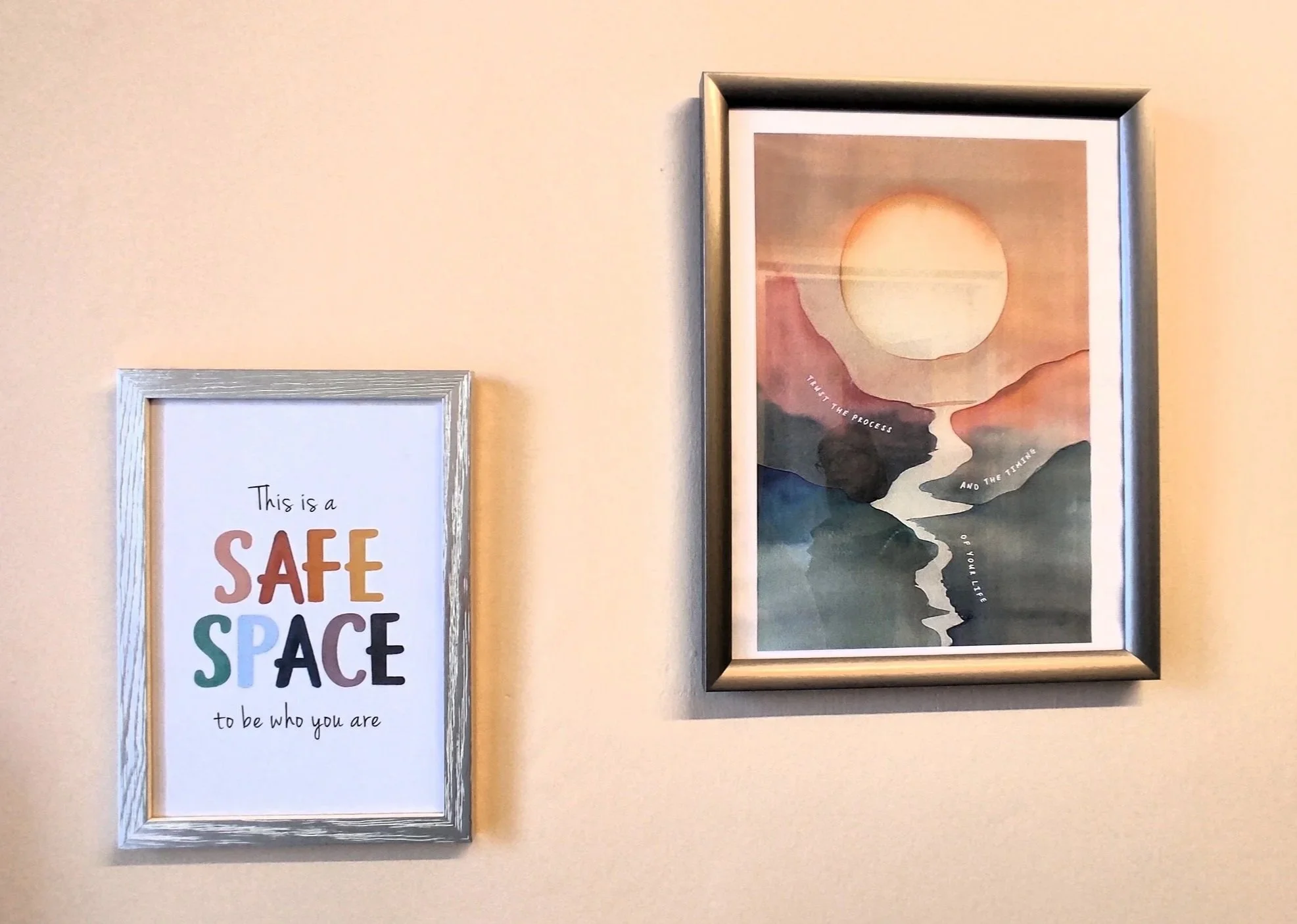 Wall with two framed pictures: one with colorful text that reads 'This is a SAFE SPACE to be who you are,' and a second with a watercolor landscape of mountains, river, and a large sun.