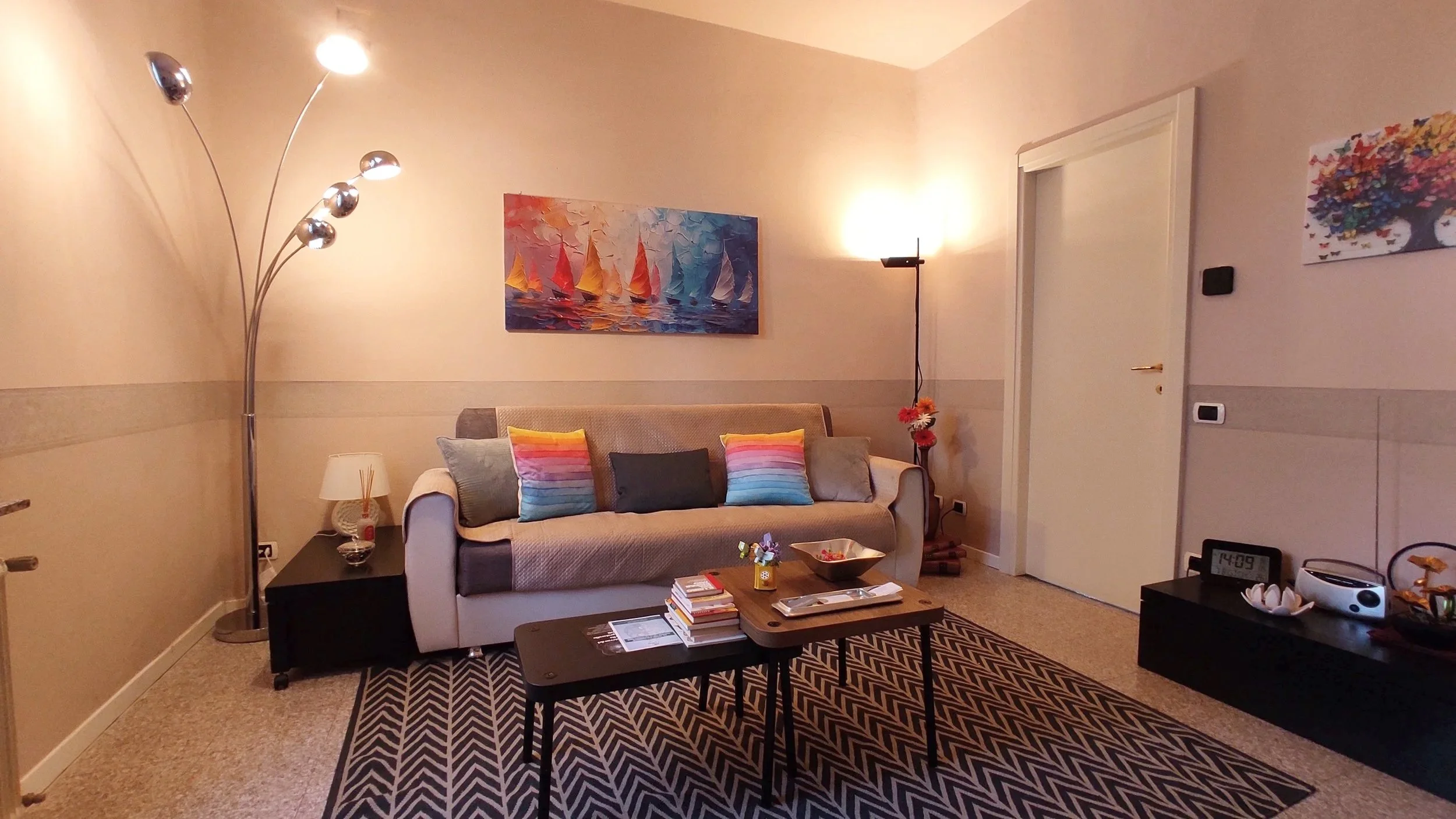 Living room with a beige sofa decorated with colorful pillows, a black side table with a lamp, a modern floor lamp with multiple curved arms, a coffee table with books and a bowl, a black entertainment unit with a clock, a stereo, and a bowl, artwork