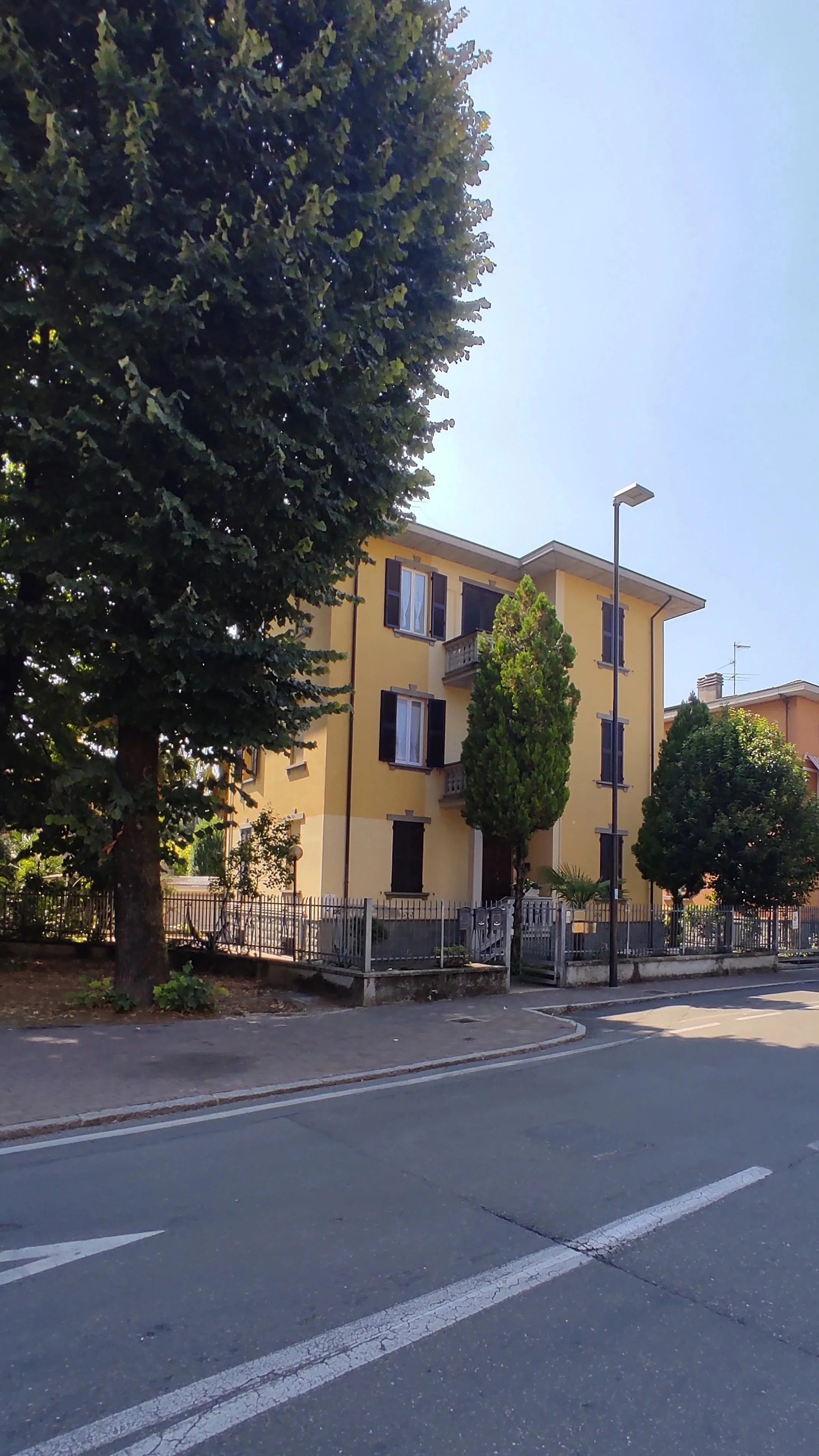A residential street with a yellow apartment building, tall trees, and a clear blue sky.