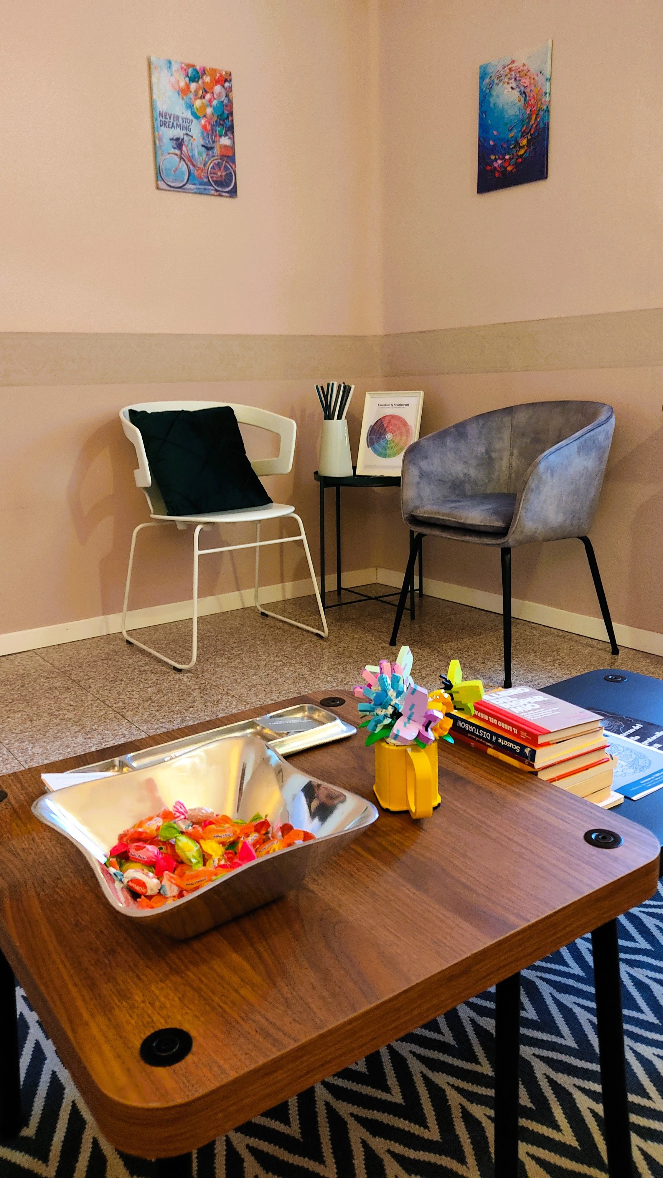 A cozy waiting room with a wooden table in the foreground holding a bowl of colorful candies, a yellow mug with pens, and a stack of books. In the background, there are two chairs—one white with a black cushion and one gray—alongside a small black si