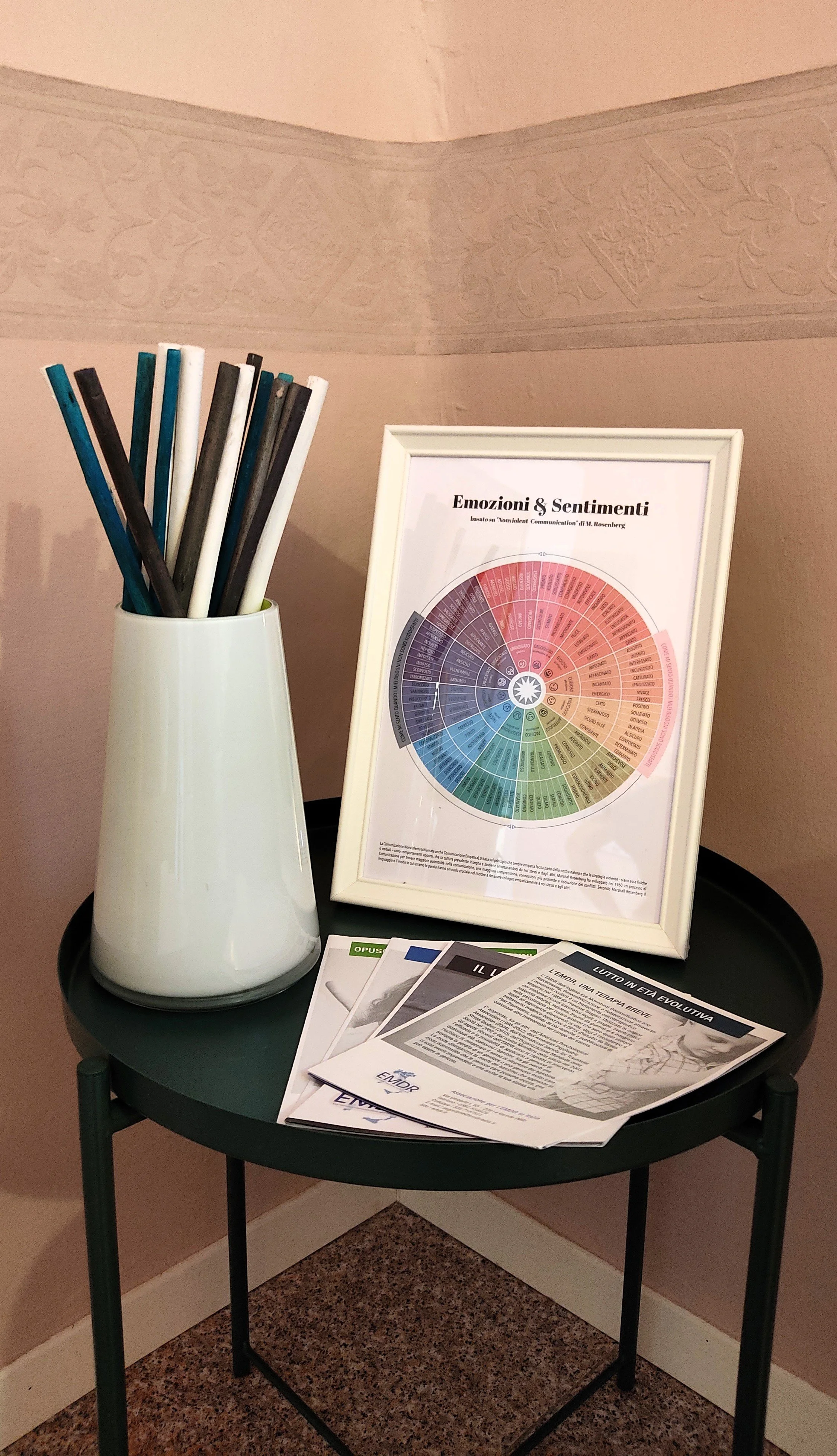 A small black round table with a white vase holding black, white, blue, and green pencils, a framed chart titled 'Emozioni & Sentimenti,' and informational brochures, set against a pink wall with decorative molding.