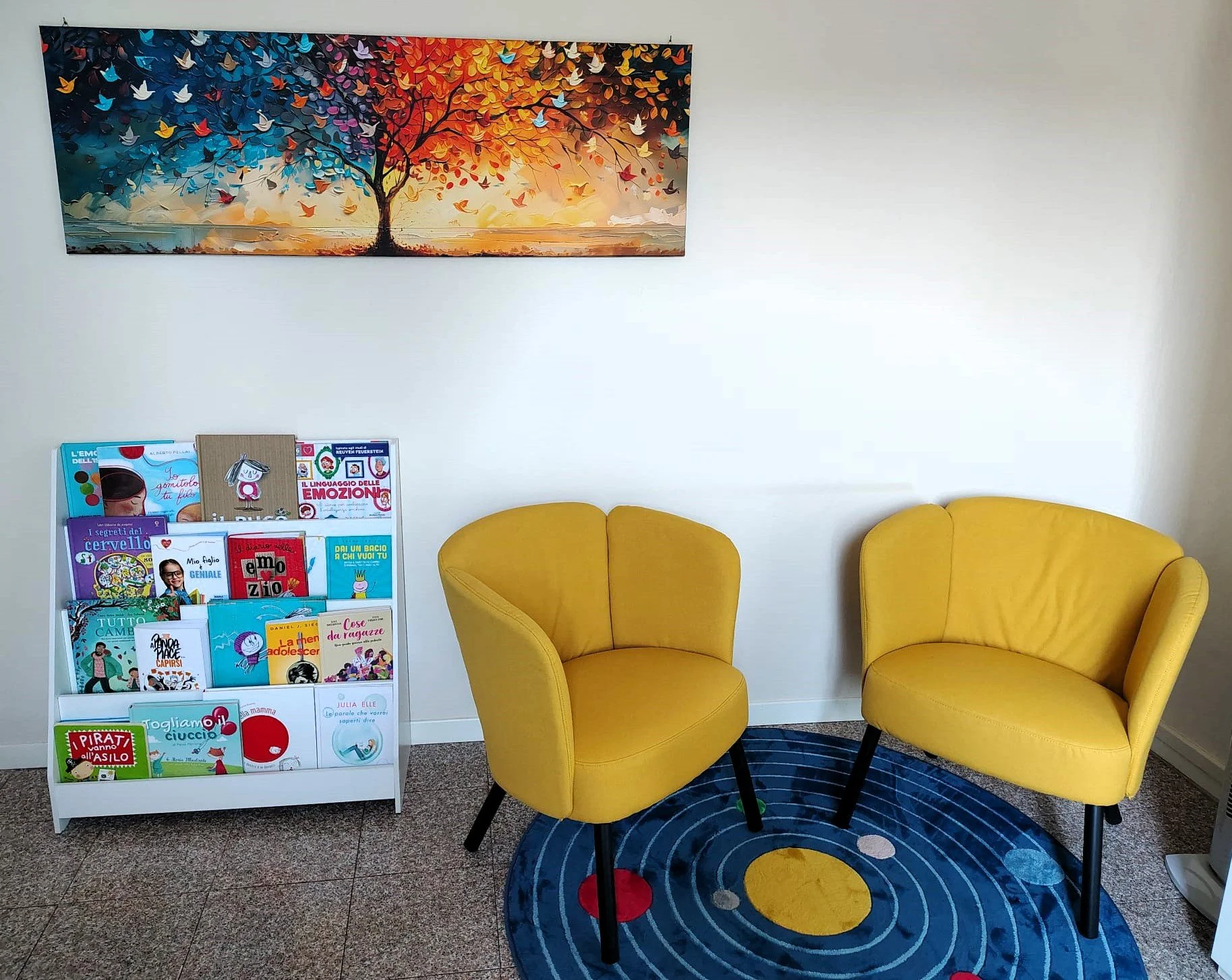 A waiting room with two yellow armchairs, a blue circular rug with yellow and red circles, a white bookshelf displaying kids' books, and a colorful painting of a tree with multicolored leaves and birds on the wall.