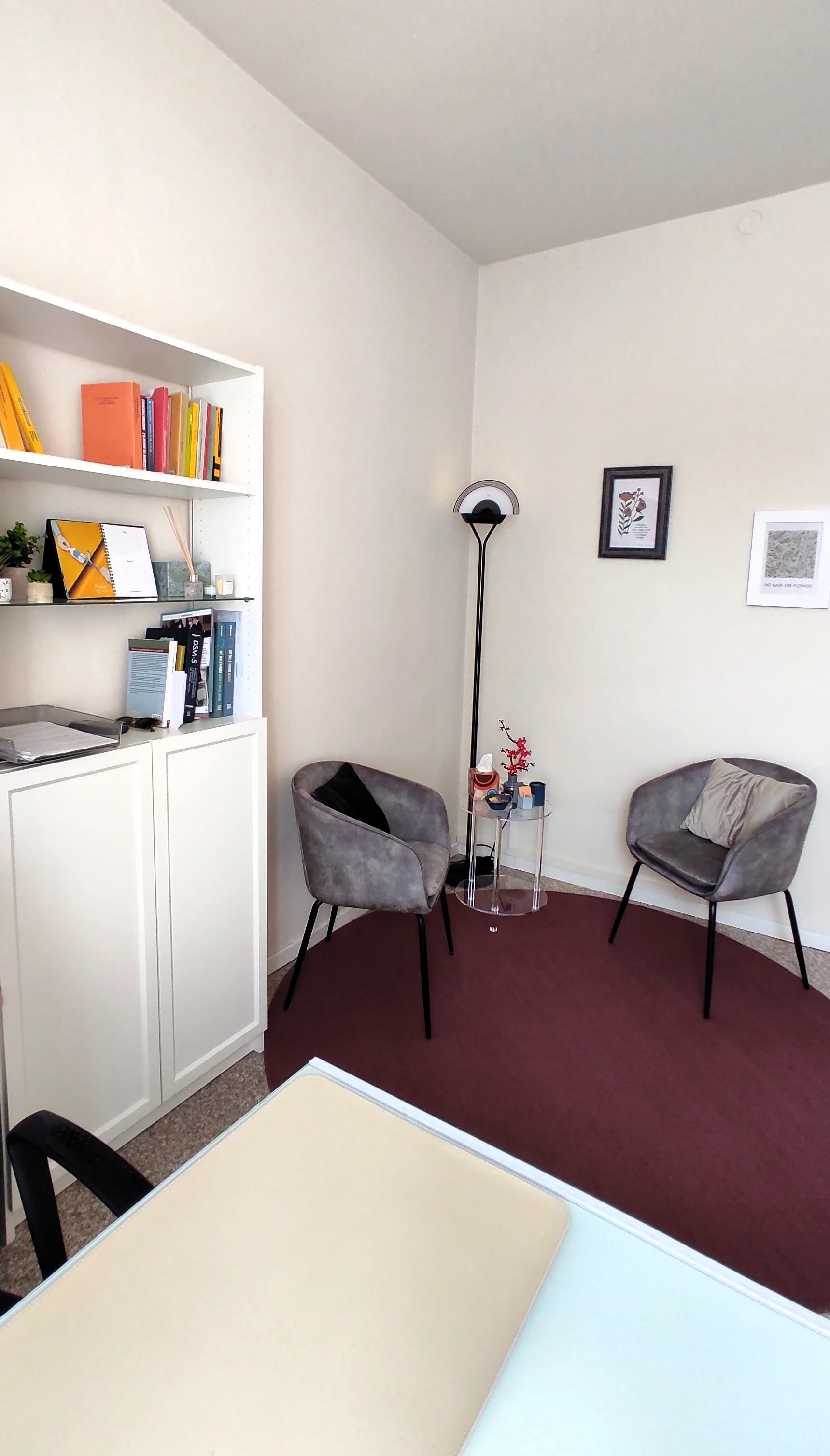 A cozy office corner with two gray chairs, a small round glass table, a white bookshelf filled with colorful books and decorative items, a floor lamp, and framed artwork on the wall.