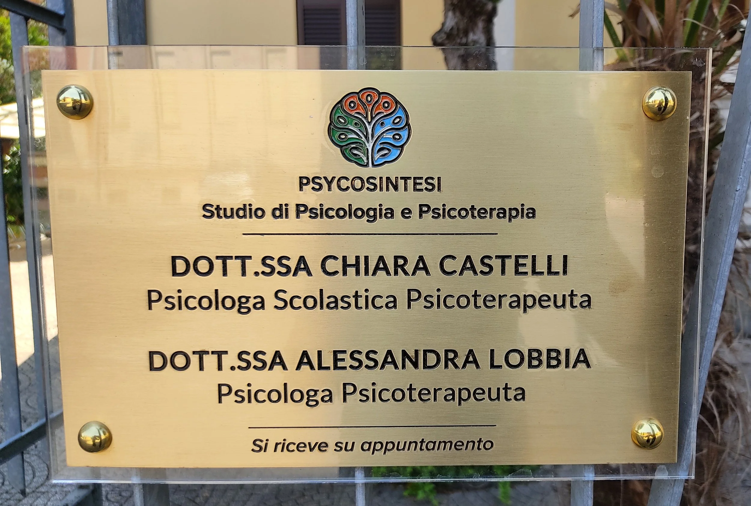 Gold-colored plaque on a fence with a logo of a colorful tree and text about psychology and psychotherapy services, including names of two psychologists, Chiara Castelli and Alessandra Lobbia, and an Italian phrase about appointments.