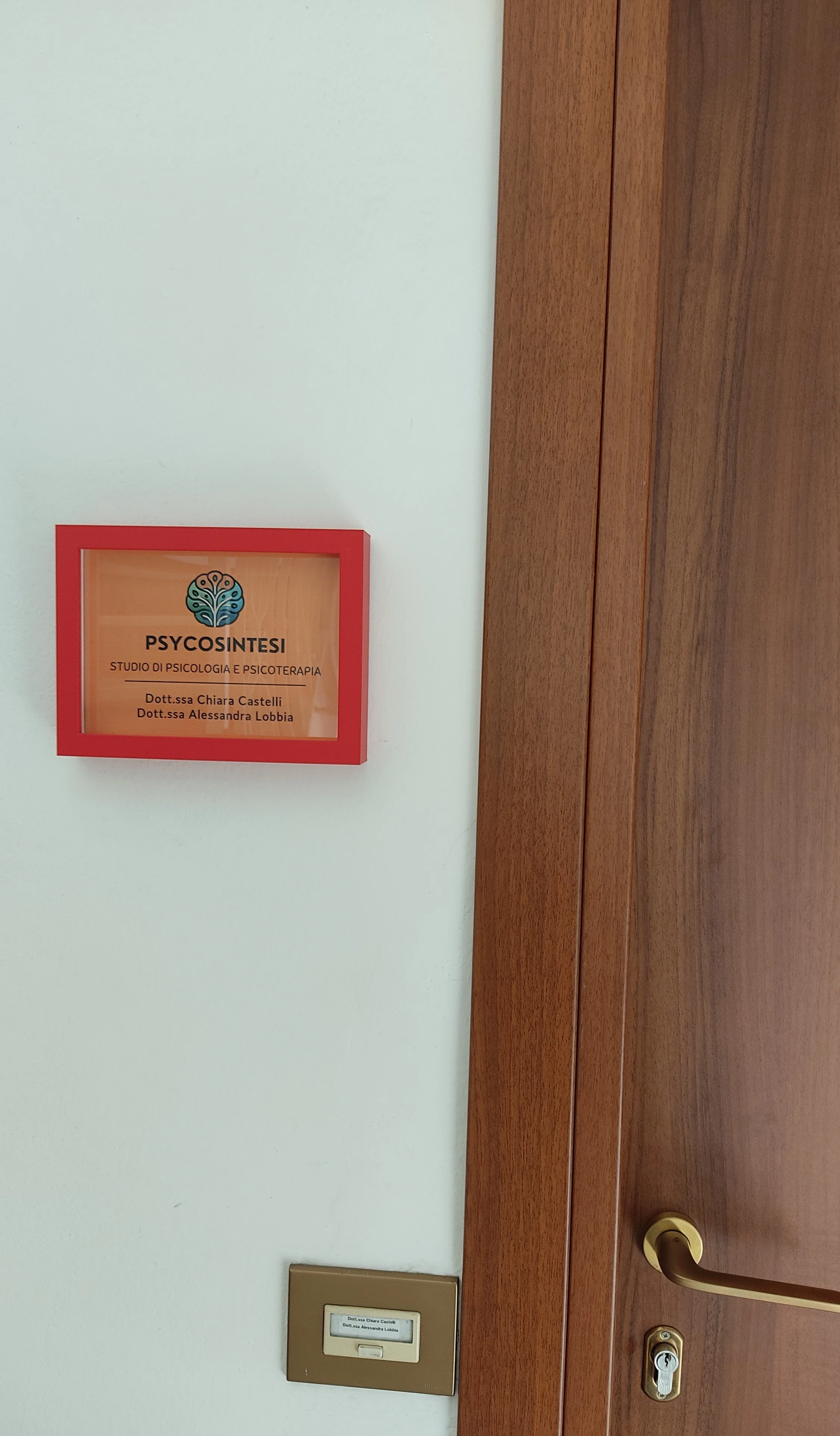 Sign on white wall reading 'PSYCOSINTESI Studio di Psicologia e Psicoterapia Dott.ssa Chiara Castelli Dott.ssa Alessandra Lobbia' with logo, next to wooden door with brass handle and lock, and a metallic nameplate below.