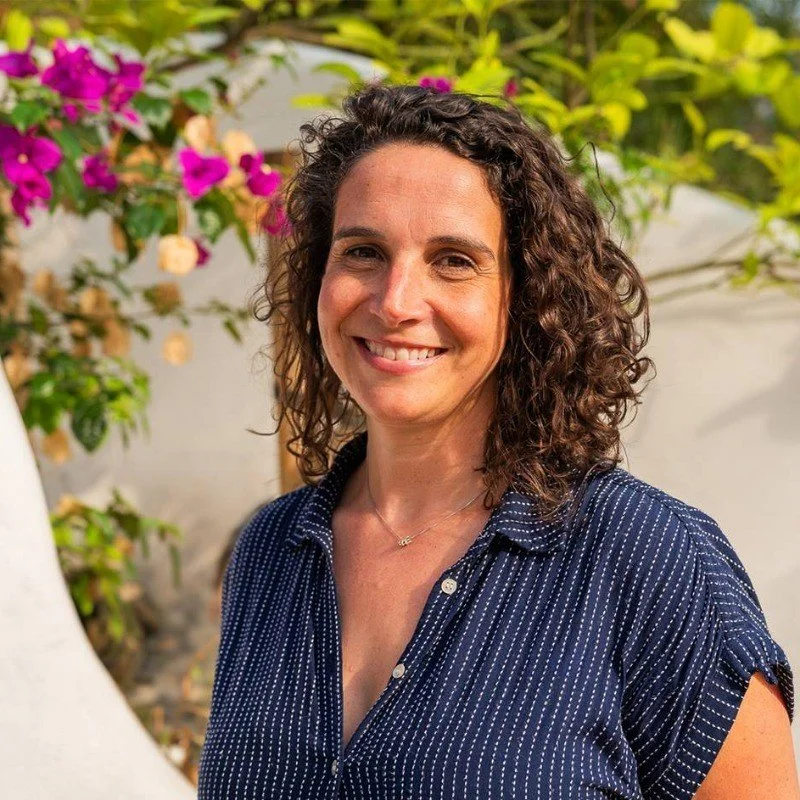 A woman with long curly hair smiles to camera with flowers in the background