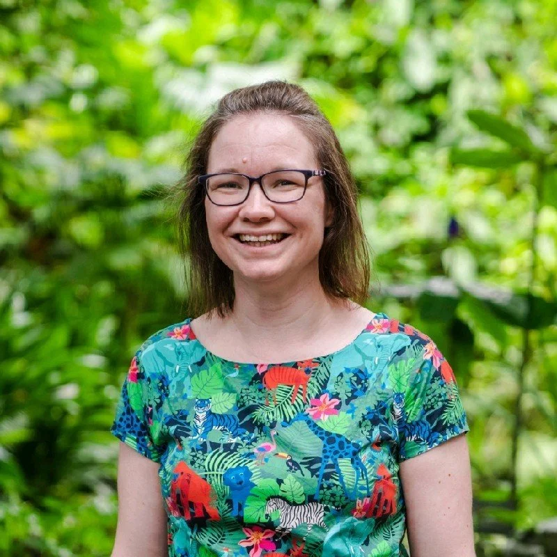 A woman with straight brown hair and glasses smiles to camera with green foliage in the background
